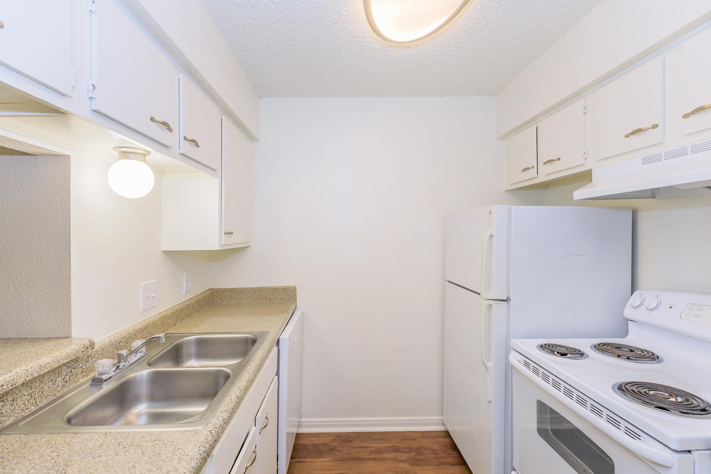 A clean and well-lit kitchen featuring white cabinets, a double sink, and a white refrigerator. There is a stove with an oven and a light fixture on the ceiling. The countertops have a speckled design, and the walls are painted a light color, giving a modern and inviting look.