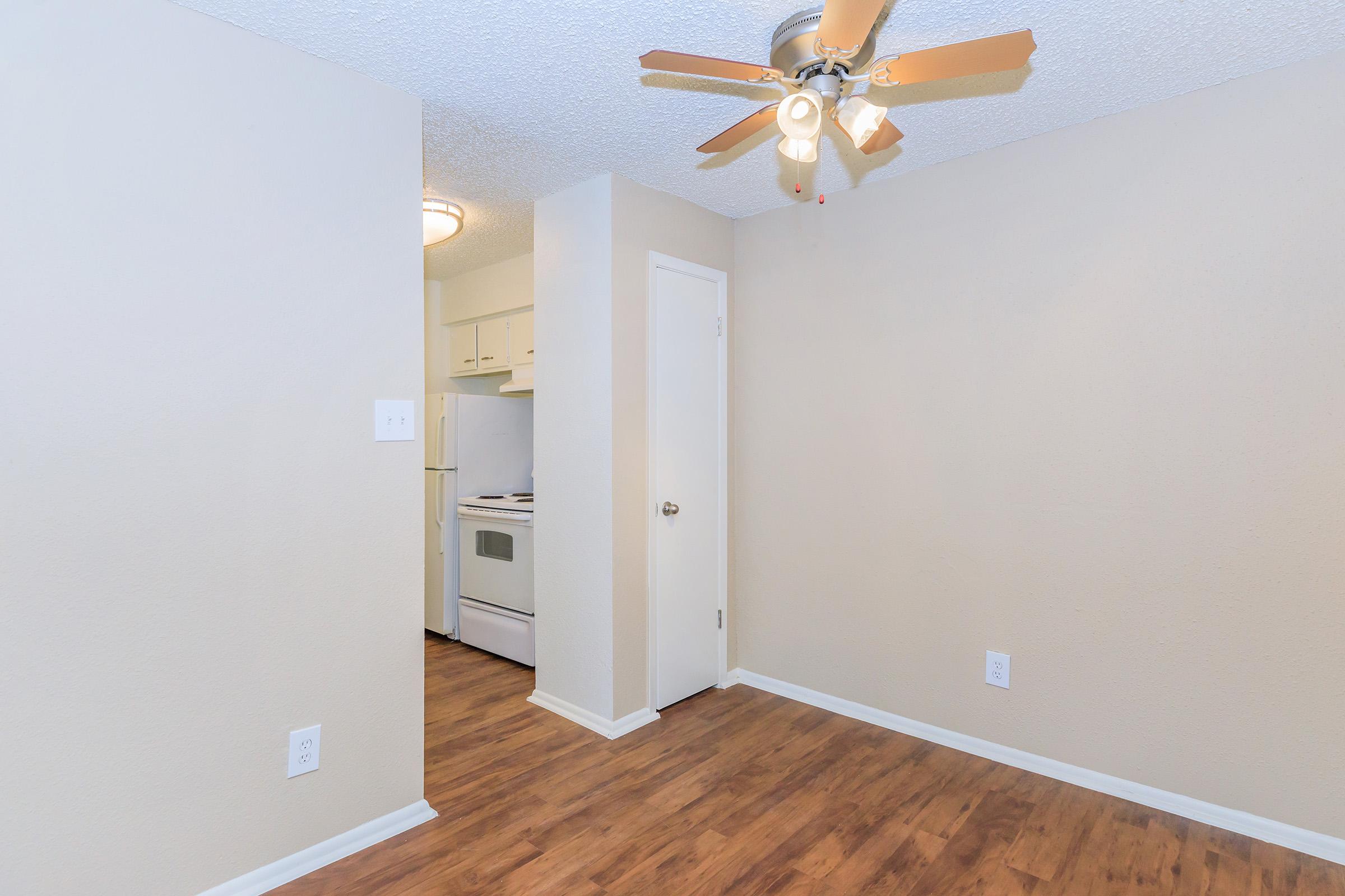 Interior view of a small room featuring a ceiling fan with light, light-colored walls, and a wood-like floor. A doorway leads to a kitchen area equipped with white appliances, including a stove and oven. The lighting is bright, creating a warm atmosphere.