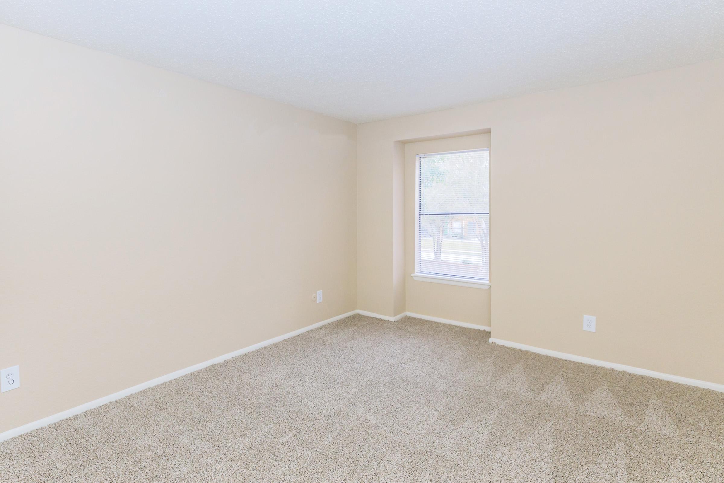 A vacant room with beige walls and carpet, featuring a window with a view. The space is empty, allowing for various layout options and furnishings. Natural light enters through the window, illuminating the room.