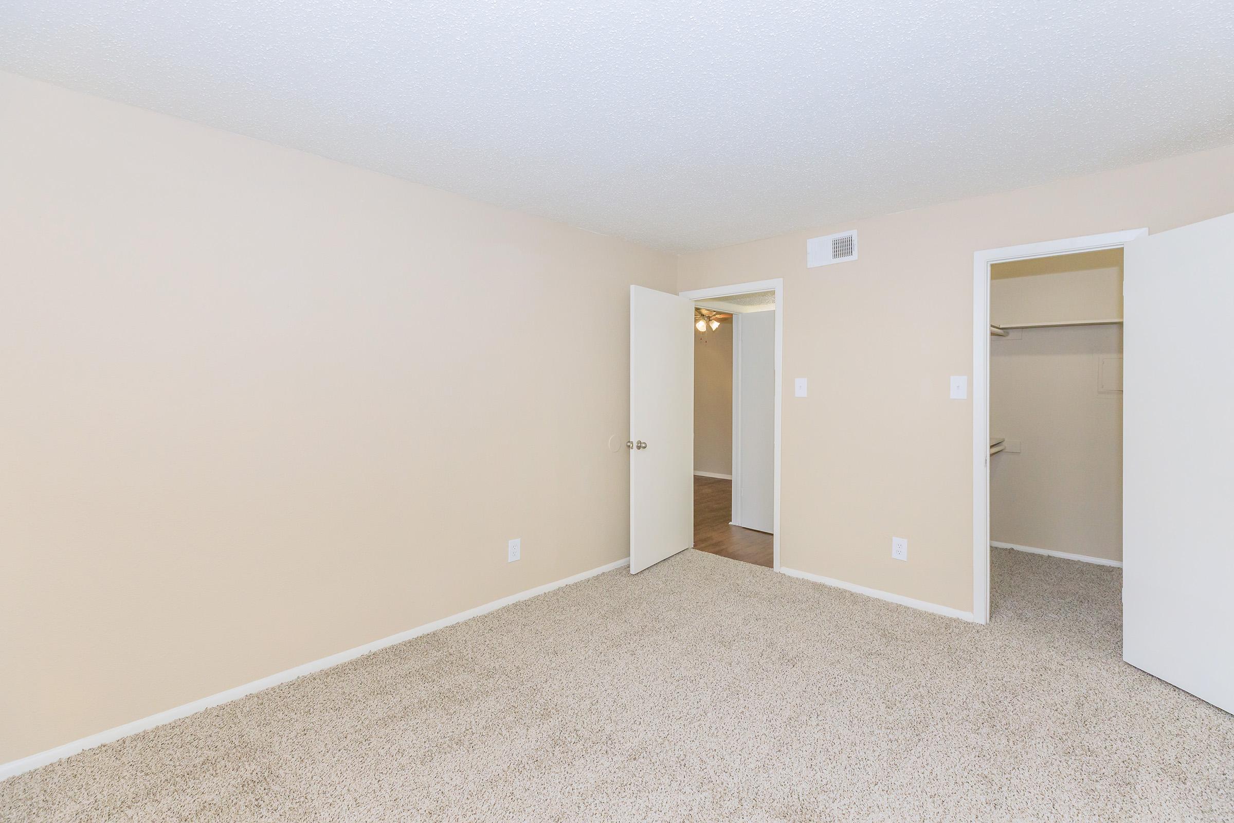 A spacious, empty room with light beige walls and carpeting. One wall features an open door leading to a closet, while another door opens to another area. The ceiling has a textured finish, and there is a light fixture visible in the adjacent space. The overall atmosphere is bright and inviting.