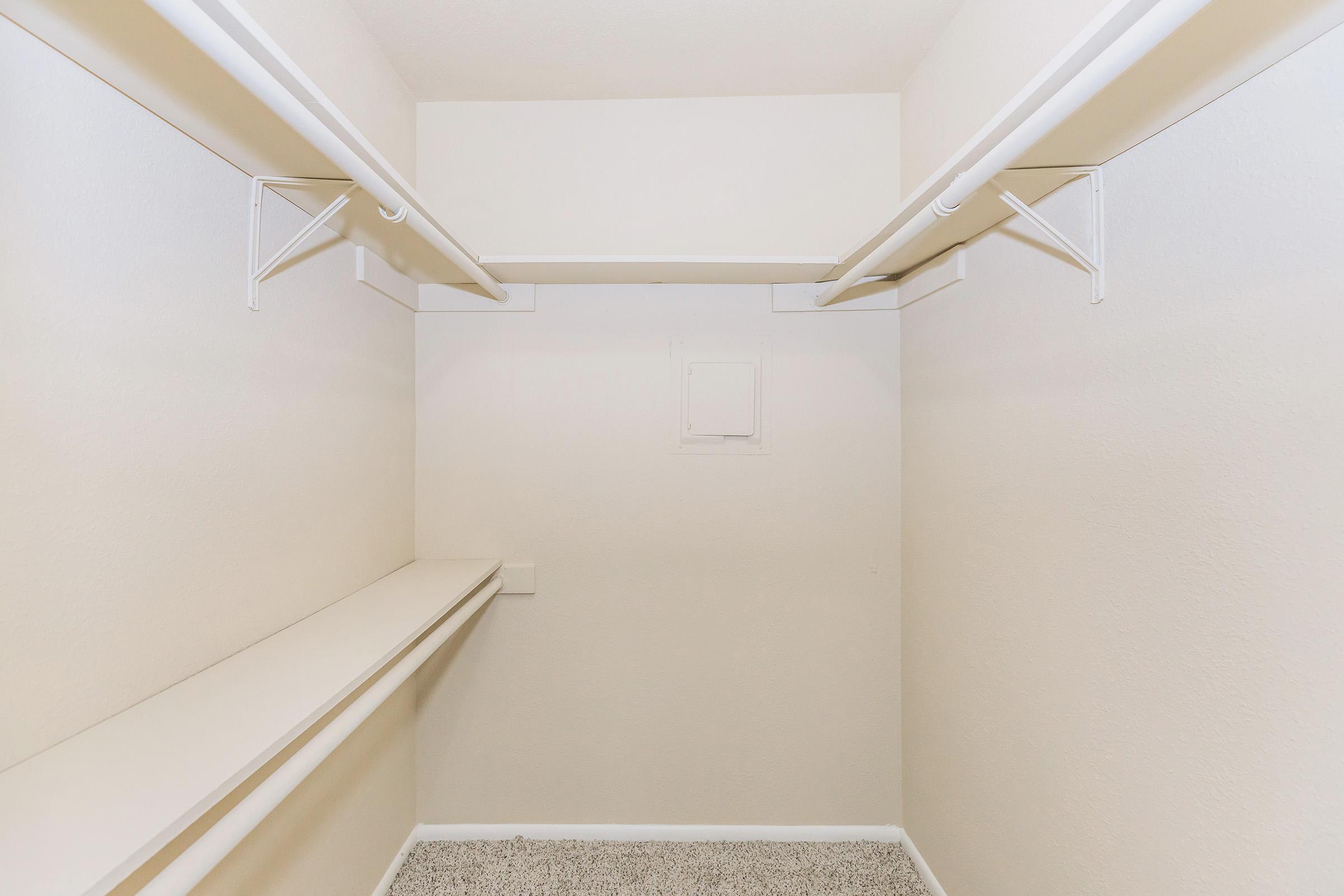 Empty closet with white walls and shelving. Two hanging rods are mounted on the walls, and there is a long shelf along one side. The floor is carpeted in a light color, and there is a small access panel on the wall near the back of the closet.