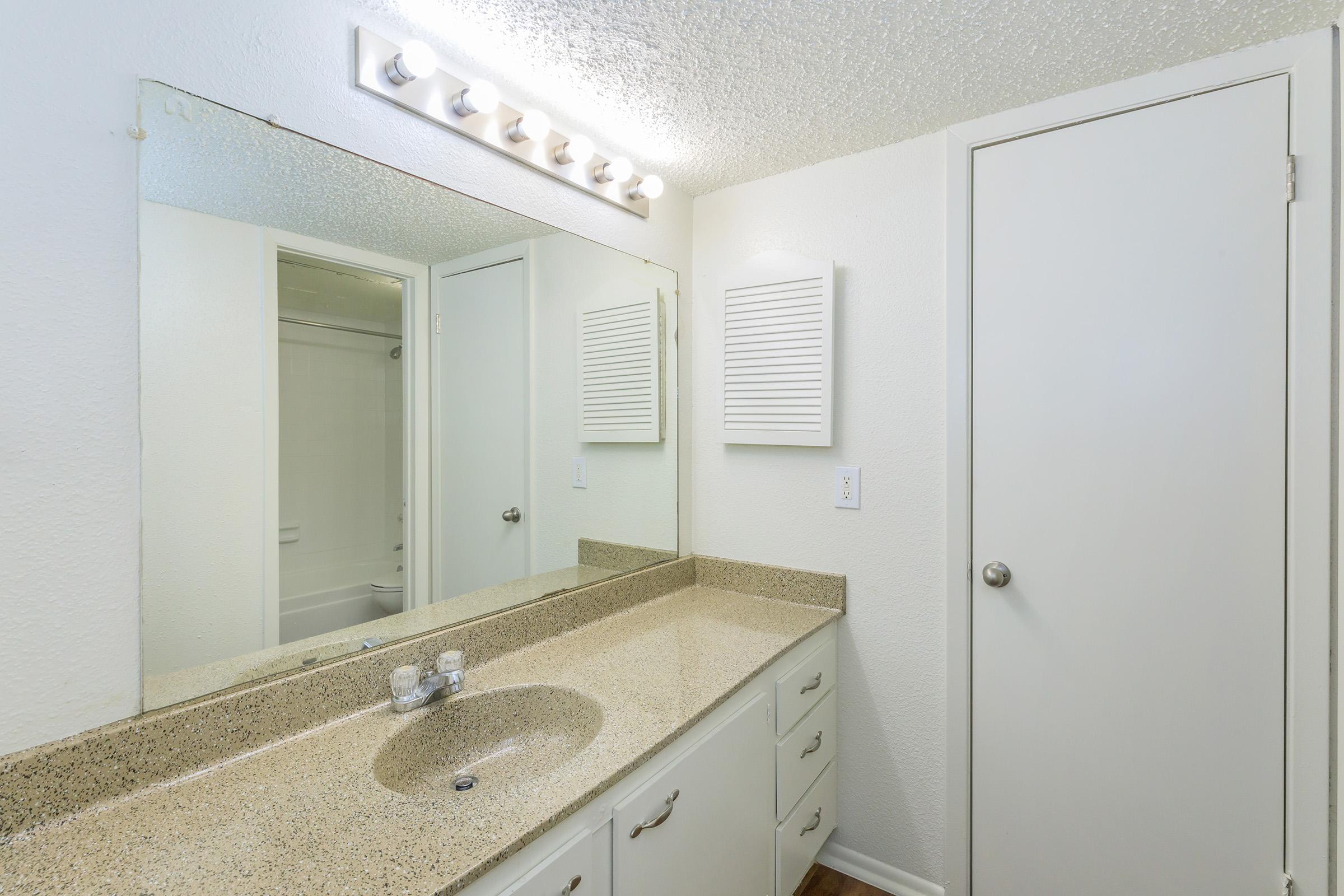 A modern bathroom featuring a granite countertop with a sink, a large mirror above the sink, and a light fixture. The walls are painted white, and there is a door leading to another area. A vent cover is mounted on the wall. The overall design is clean and neutral, with a bright and spacious feel.