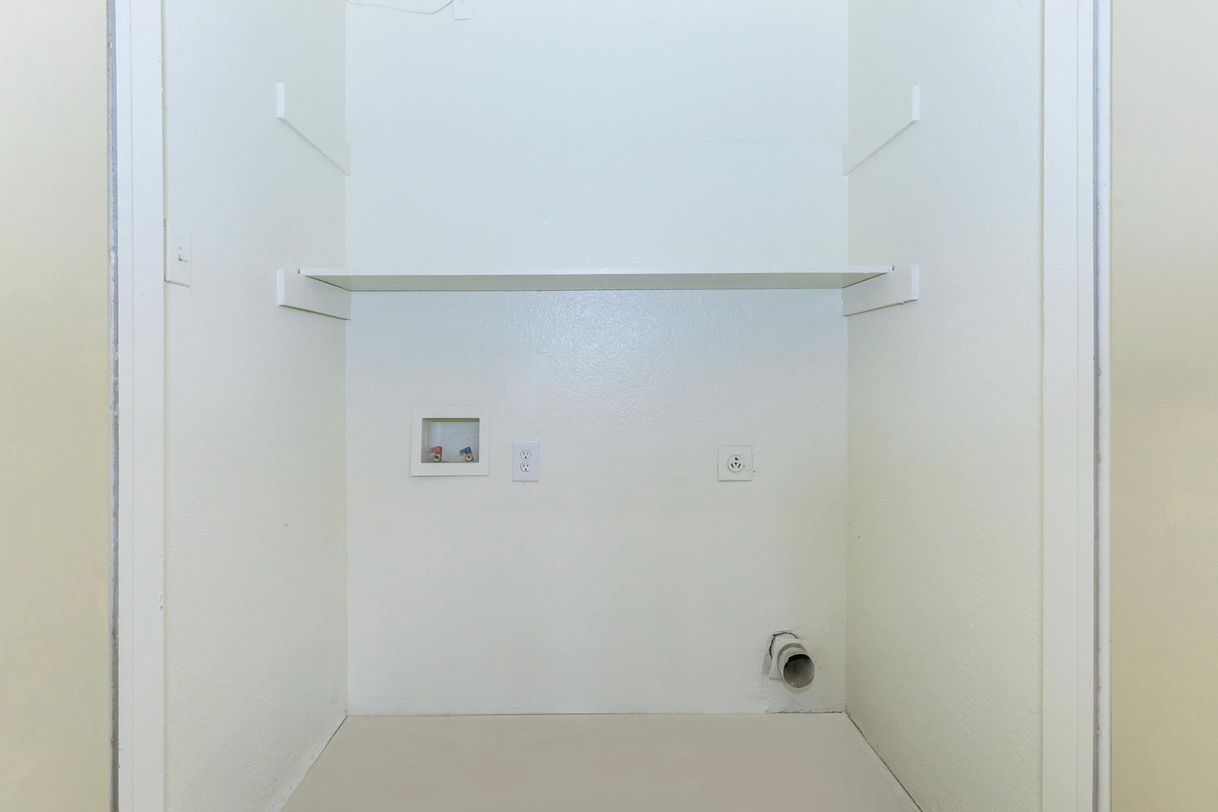 Empty room with plain white walls and a concrete floor. A single shelf is mounted on the wall, and there are two electrical outlets and a small vent or pipe at the bottom. The space appears to be an unfinished area, possibly a closet or utility room, with no furniture or decorations.