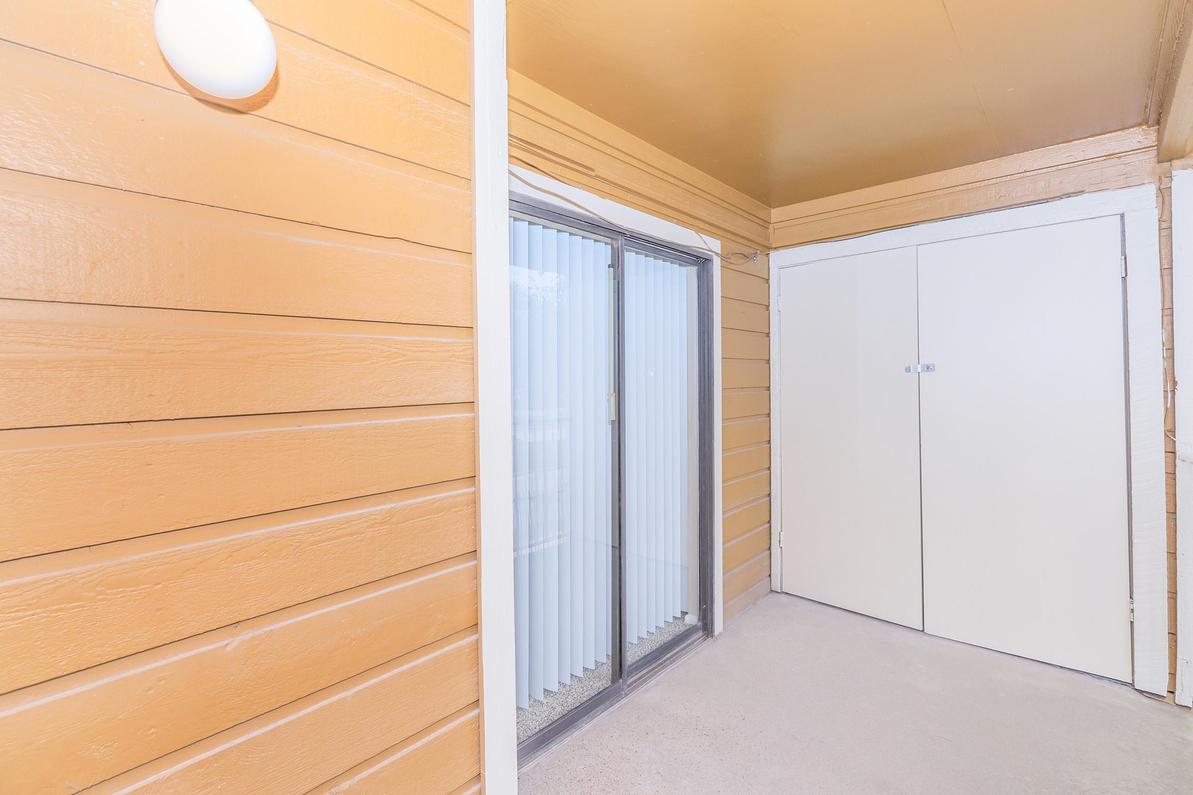 A covered entryway featuring wooden paneling in an orange hue, with a sliding glass door to the left and a closed storage closet door on the right. The floor is concrete, and the ceiling is painted in a light color. Soft lighting is provided by a wall-mounted fixture.