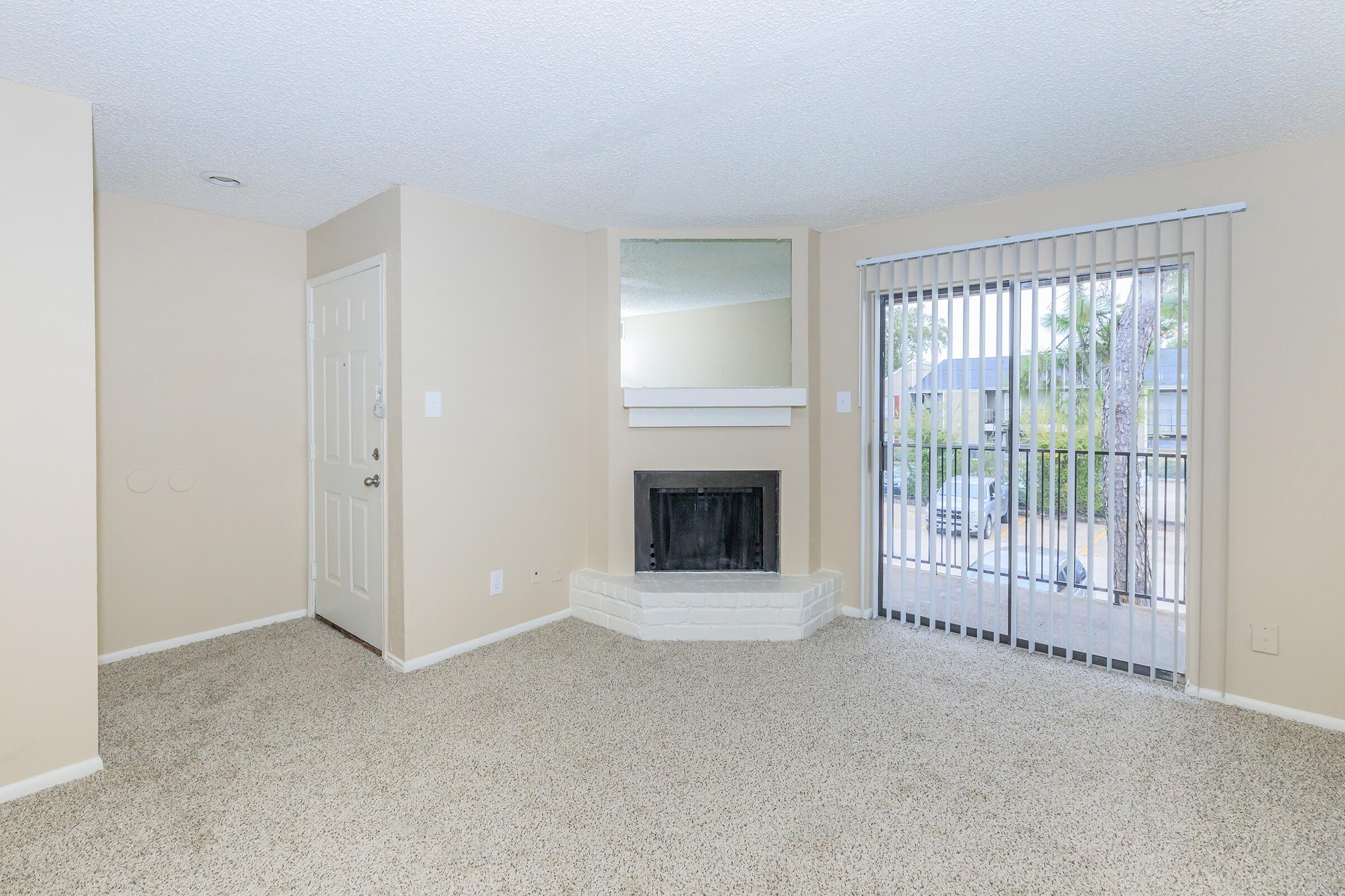 A cozy living room featuring beige walls and carpet. It includes a fireplace with a white mantel, a large sliding glass door with vertical blinds leading outside, and a small entryway with a white door. Natural light enters the room, creating a warm atmosphere.