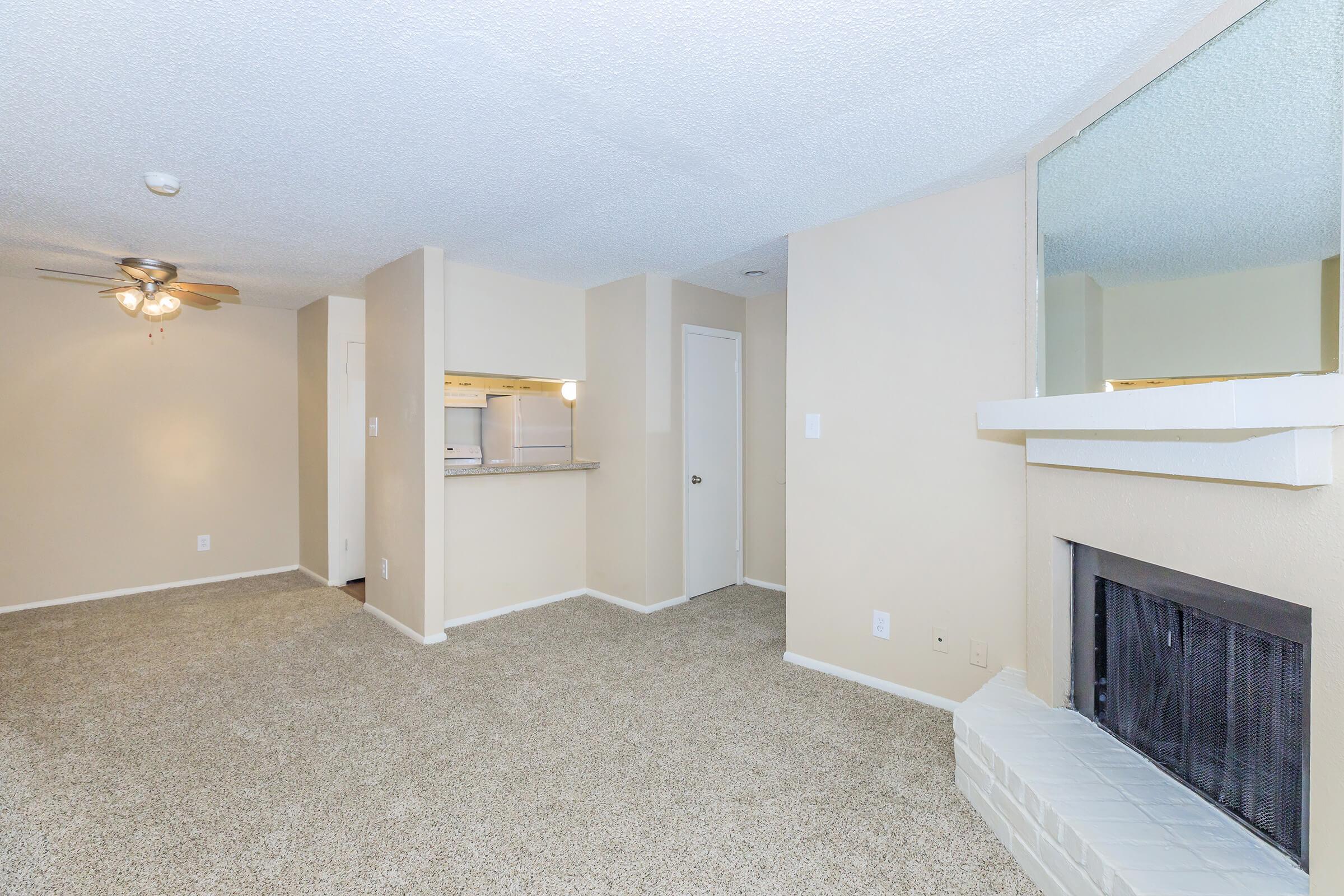 Spacious living area featuring a ceiling fan, light-colored walls, and plush carpet. A partial view of the kitchen with a bar counter is visible on the left, and a cozy fireplace with a white mantel is situated on the right side. Natural light reflects off the walls, creating a warm atmosphere.