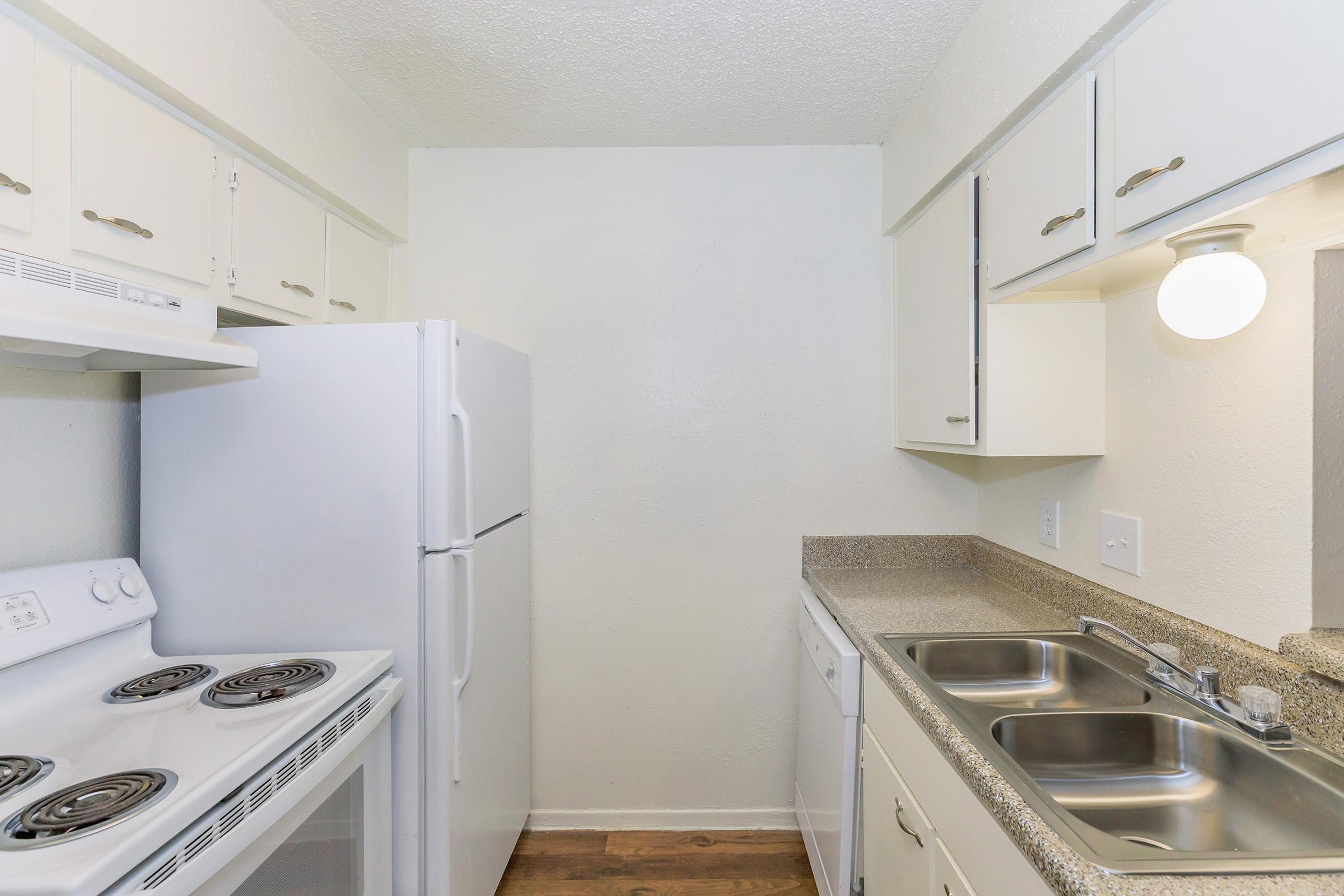 A small kitchen featuring white cabinets, a white refrigerator, a stove with burners, and a dual sink. The countertop is beige with a textured finish, and the walls are painted light colors, creating a bright and clean atmosphere. A round light fixture is mounted on the wall above the sink.