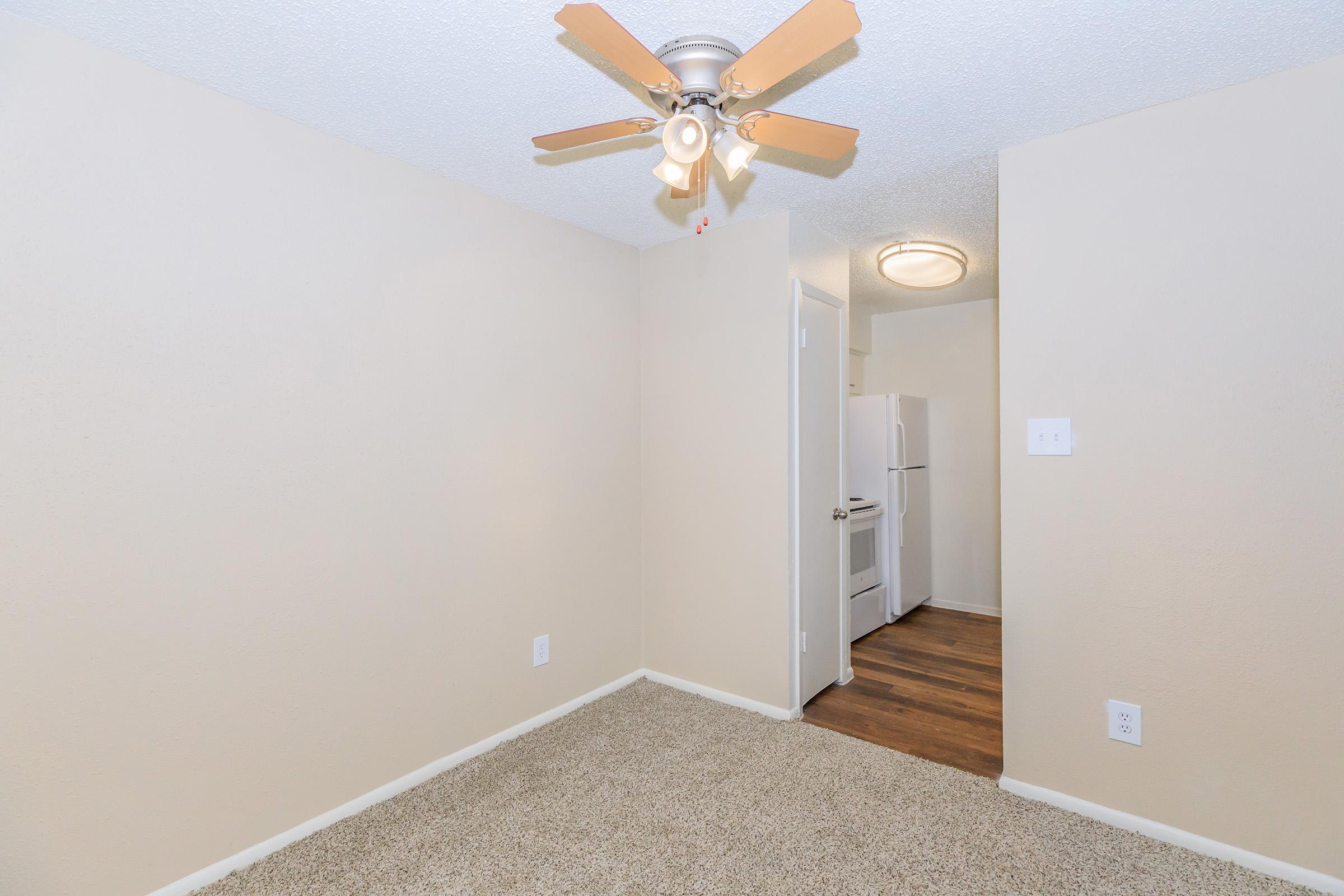 A small, empty room with a ceiling fan and beige walls. The floor is covered in carpet, and there is a door leading to another area. A kitchen space is visible in the background, featuring a white refrigerator. The overall ambiance is neutral and inviting.