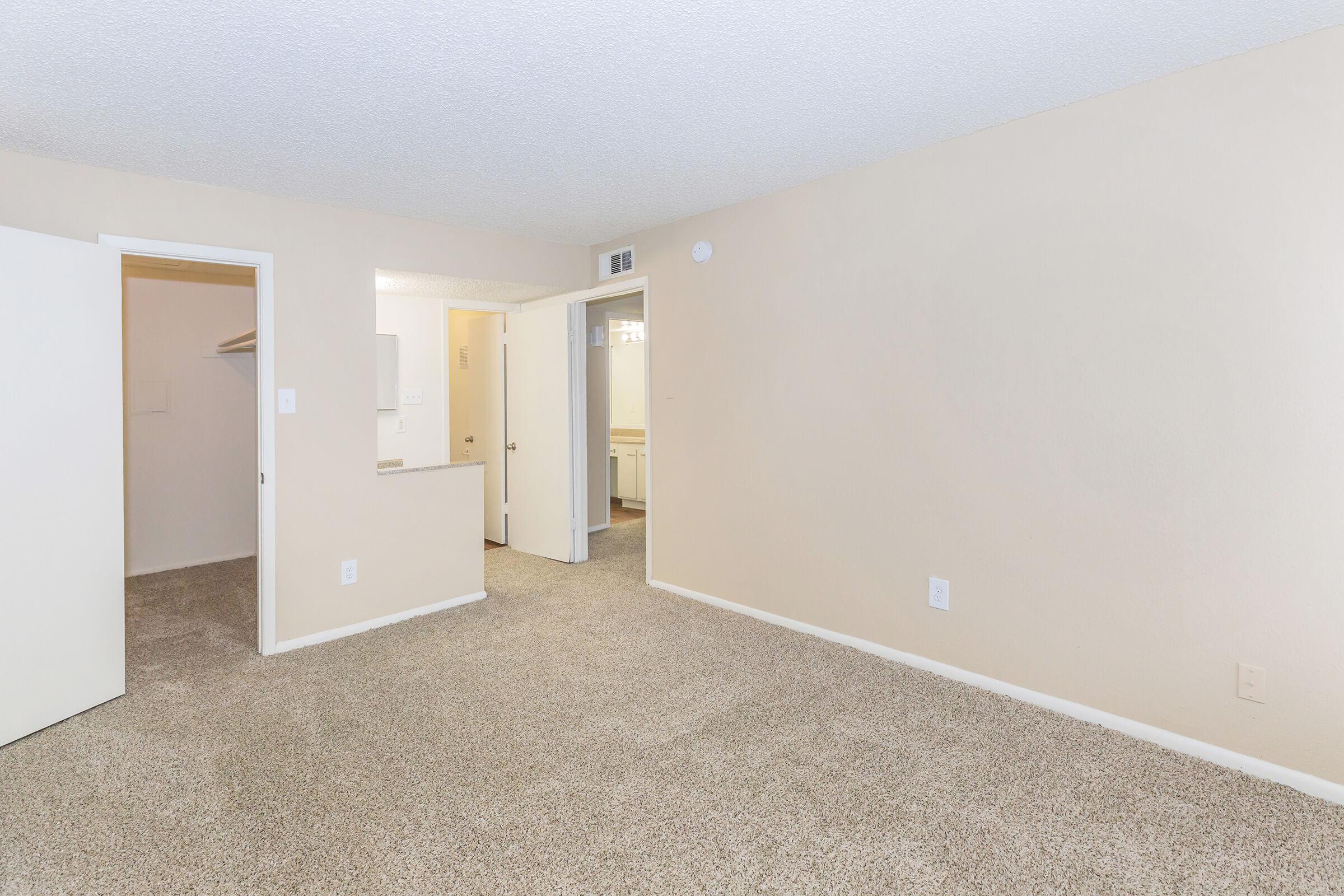 A spacious, empty room with light beige walls and carpeted flooring. One wall features a closed white door, while an open doorway leads to another area. The overall ambiance is bright and neutral, suitable for various uses such as a living space or office.