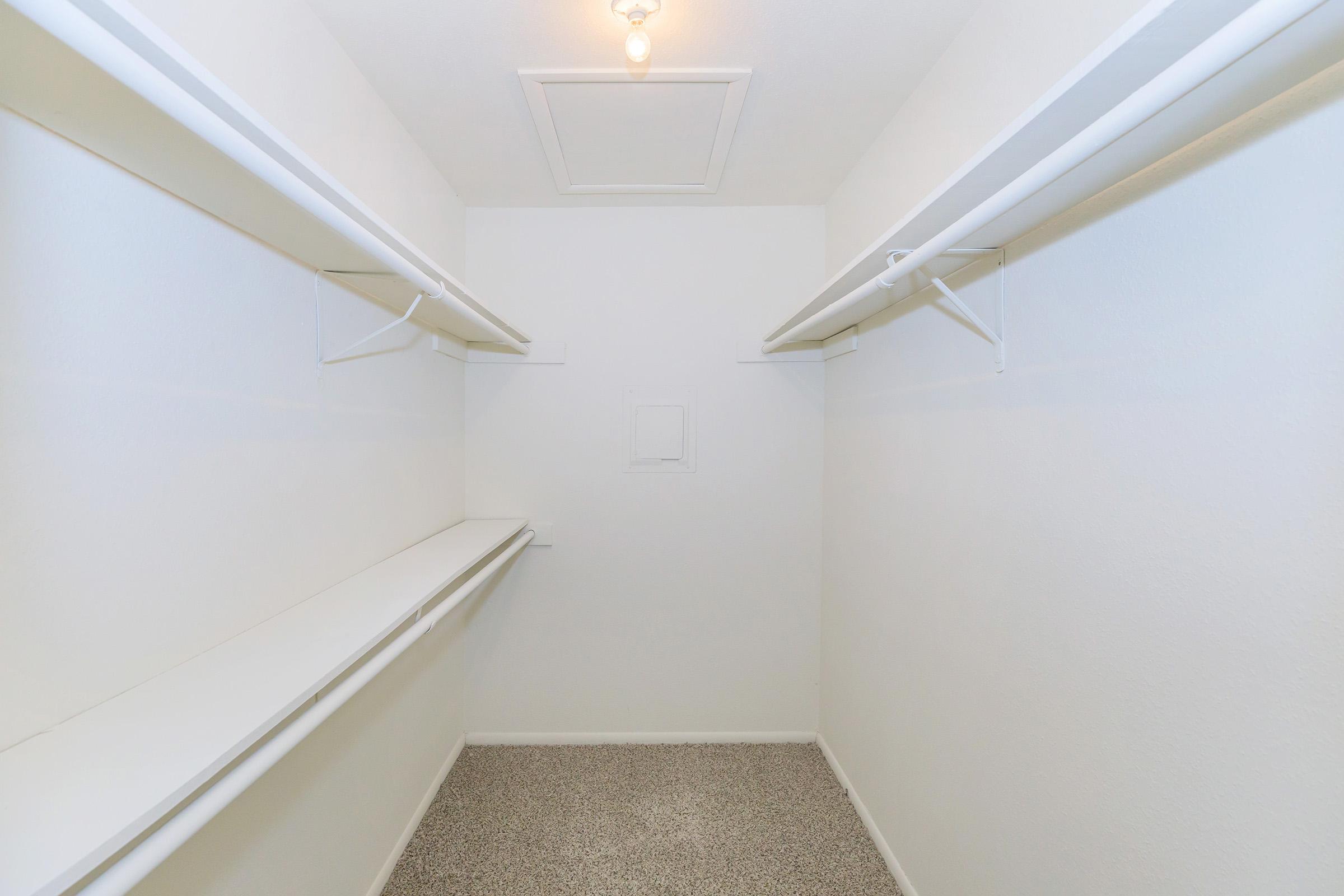 Empty closet with white walls and a light fixture. Two parallel shelves run along each side, providing storage space. The floor is covered in gray speckled material, and there's a small access panel on the ceiling. The space is clear and uncluttered, creating a minimalist appearance.