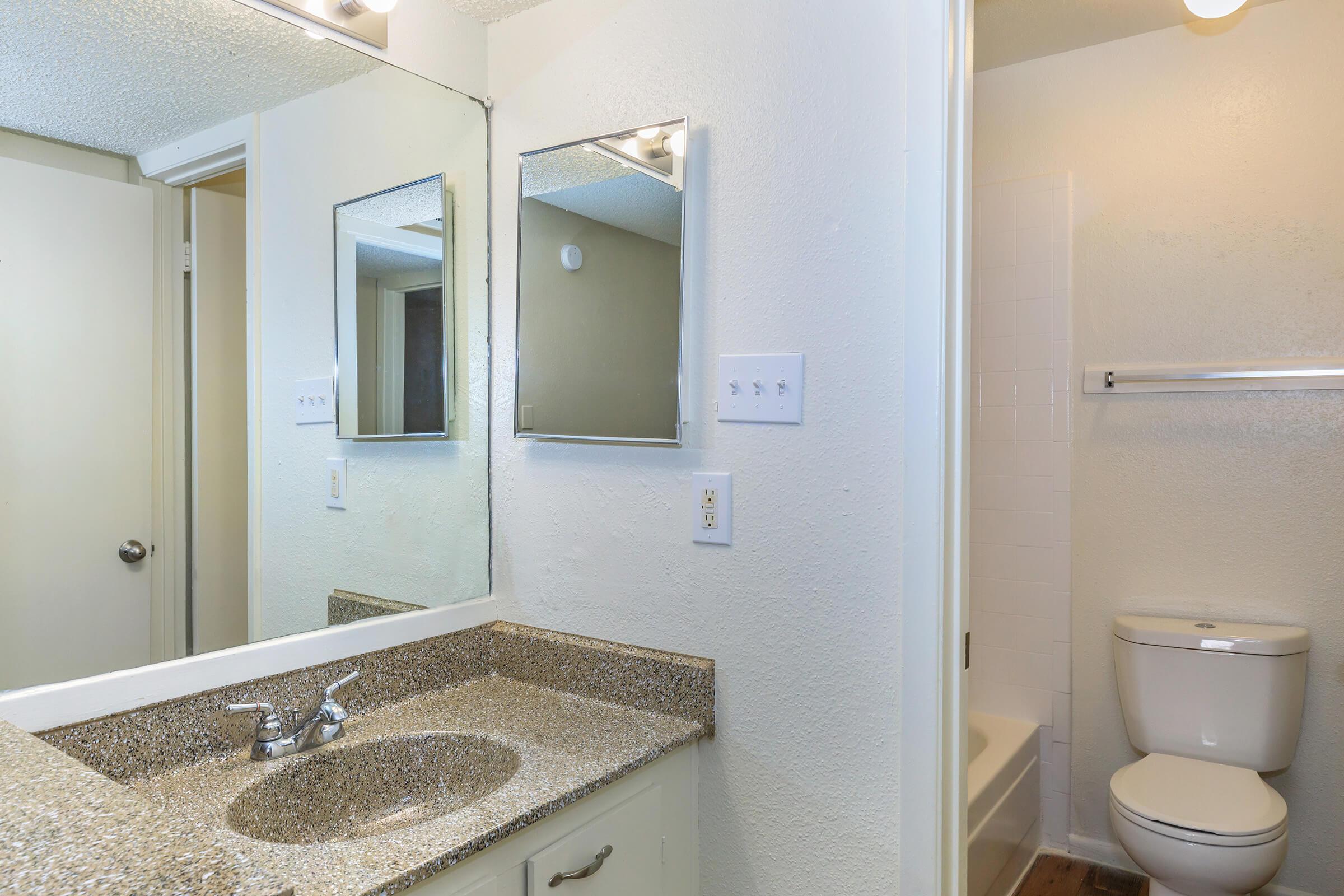 A well-lit bathroom featuring a mirror above a granite countertop with a sink, beige walls, and a white toilet. The room has a shower/tub combination visible in the background. The overall decor is simple and modern, with natural light enhancing the space.