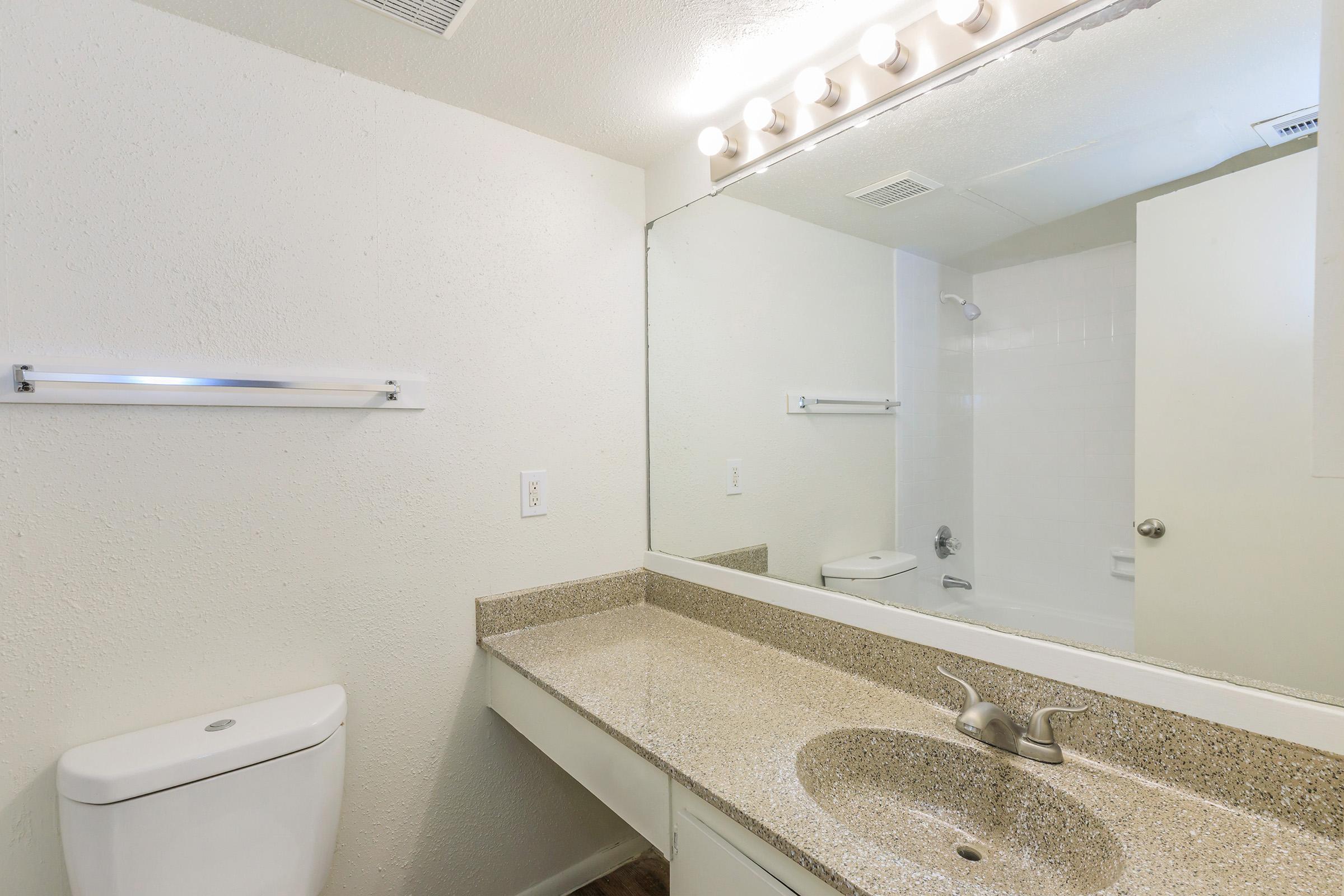 A clean, modern bathroom featuring a white toilet, a long vanity with a light-colored countertop, and a large mirror above the sink. The space has neutral-colored walls and a shower area visible in the background, with bright lighting from fixtures above the mirror.