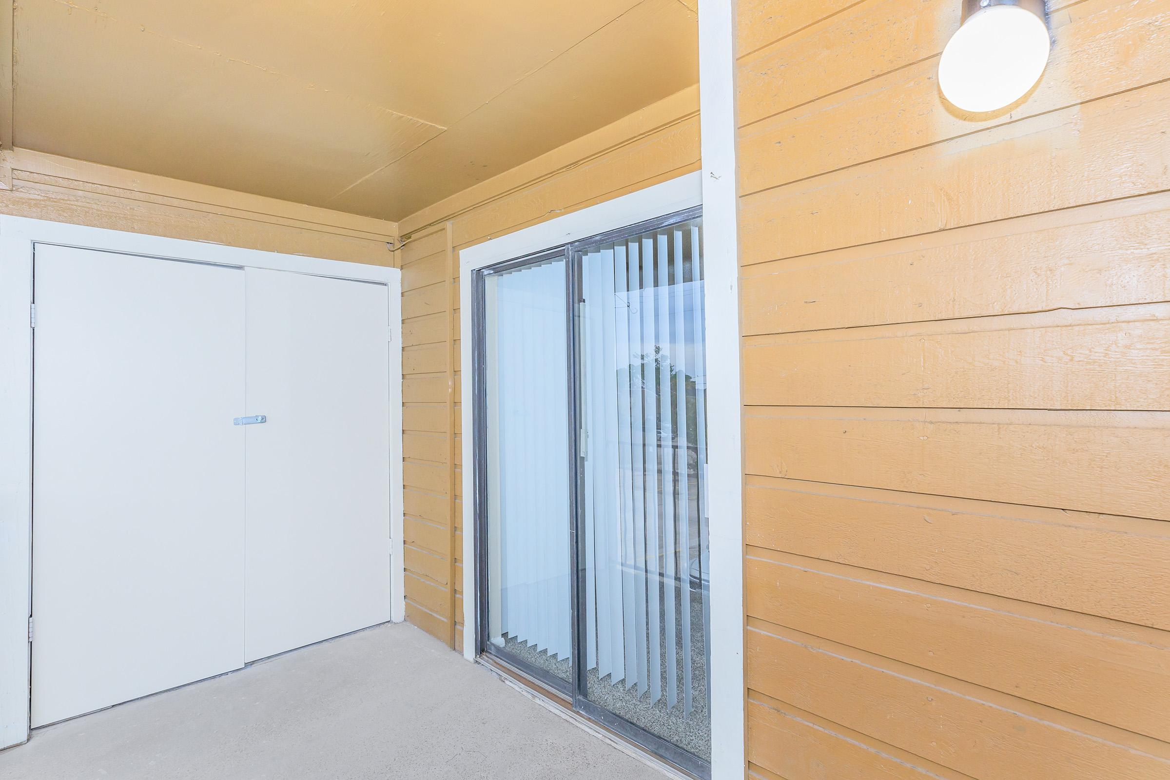 A well-lit entryway featuring a sliding glass door with vertical blinds, adjacent to a white panel door. The walls are painted a warm wood color, and a light fixture is mounted on the ceiling, creating an inviting atmosphere. The outdoor view is partially visible through the glass door.