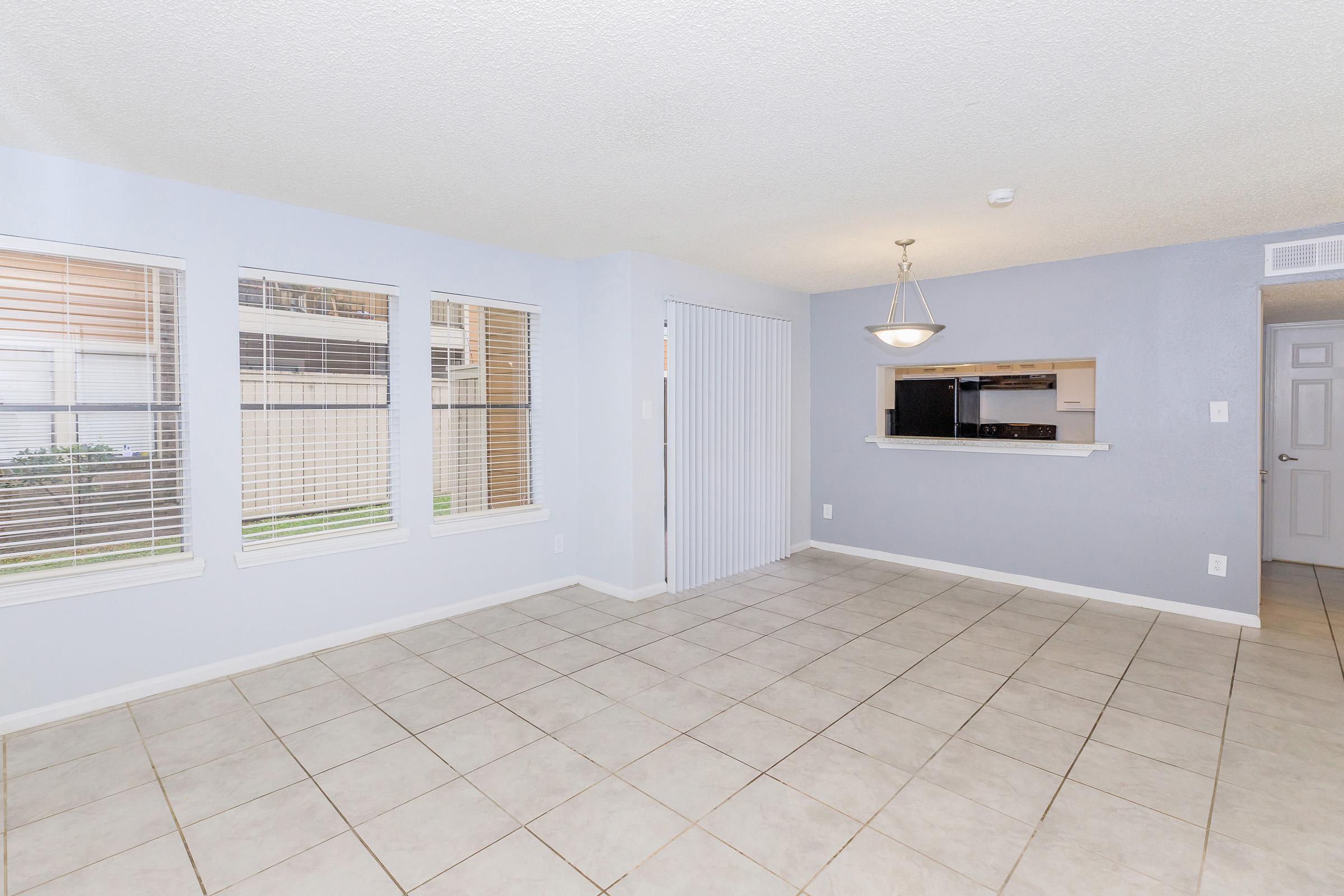 Bright and spacious living area with tiled flooring, featuring three windows allowing natural light, a sliding door to the outside, and an open layout that connects to a kitchen area with a breakfast bar. The walls are painted in soft blue tones, enhancing the airy feel of the space.