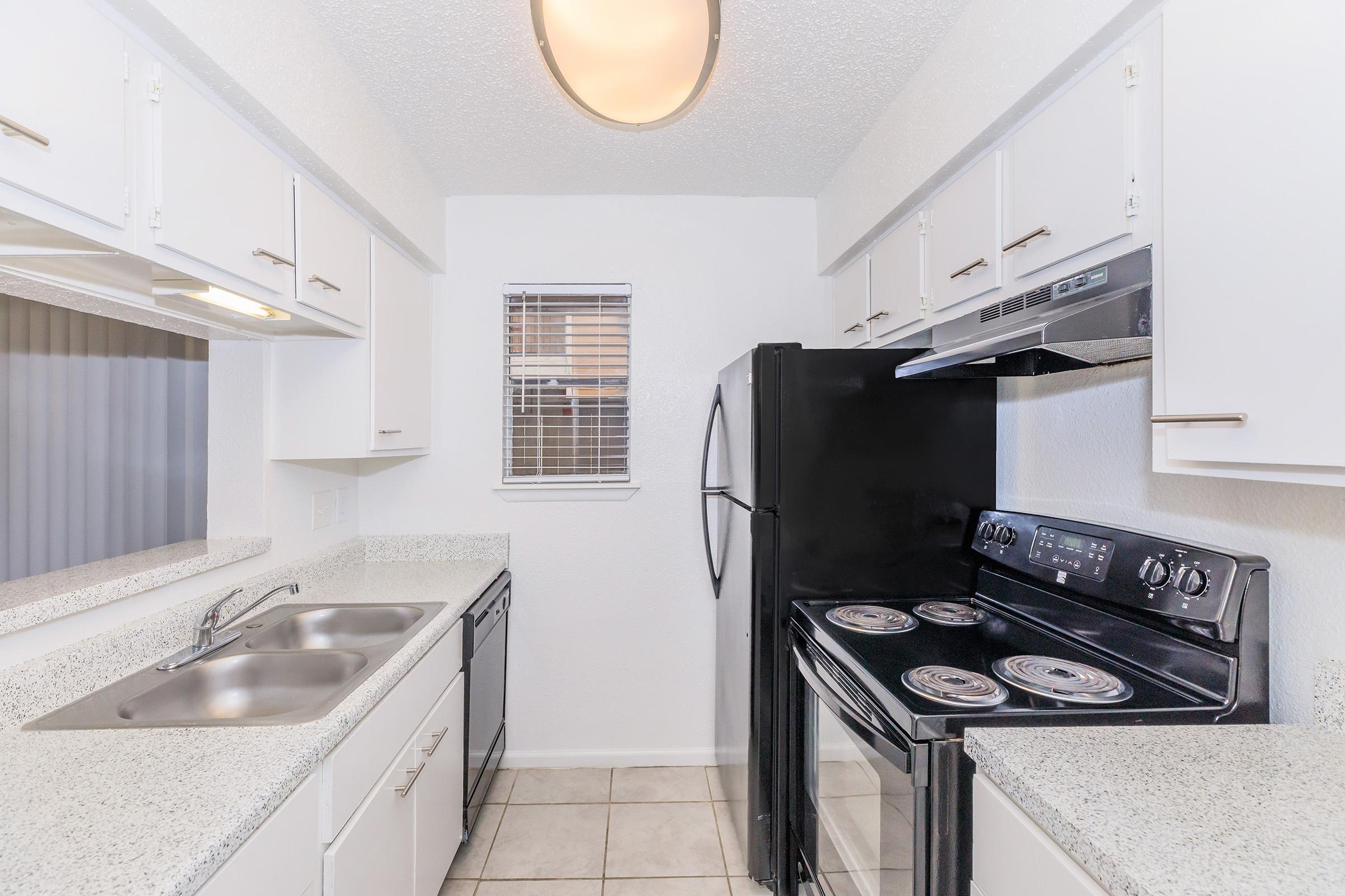 A modern kitchen featuring white cabinetry, a double sink, a black refrigerator, and a black electric stove with four burners. The countertops are light granite, and the walls are painted white. There is a window with blinds allowing natural light in. The flooring is tiled.