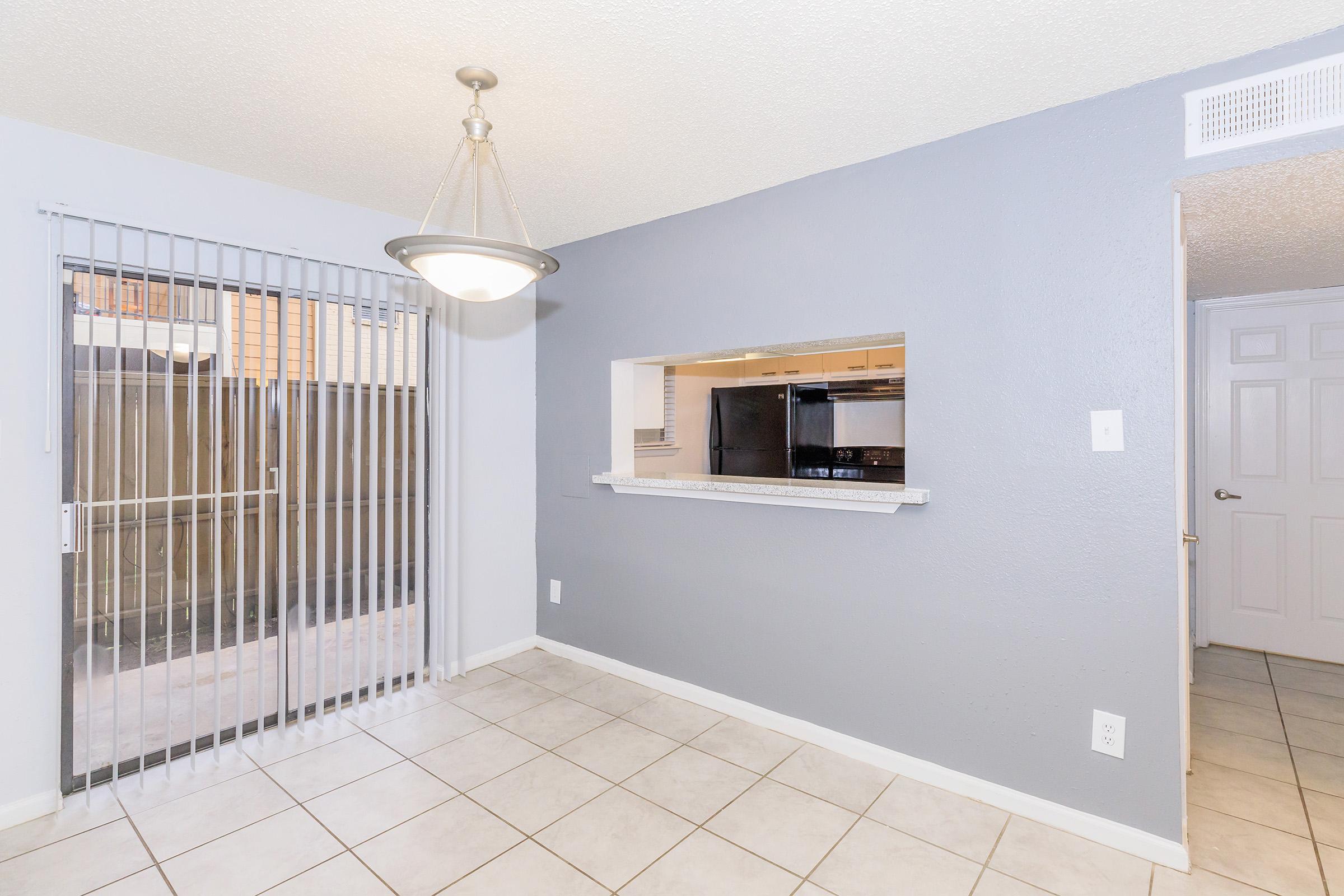 A bright dining area featuring tiled flooring, a modern light fixture hanging from the ceiling, and a large window with vertical blinds. There is a pass-through to a kitchen area on the left, which has dark cabinetry. A door on the right leads to another room, and the walls are painted in light gray.