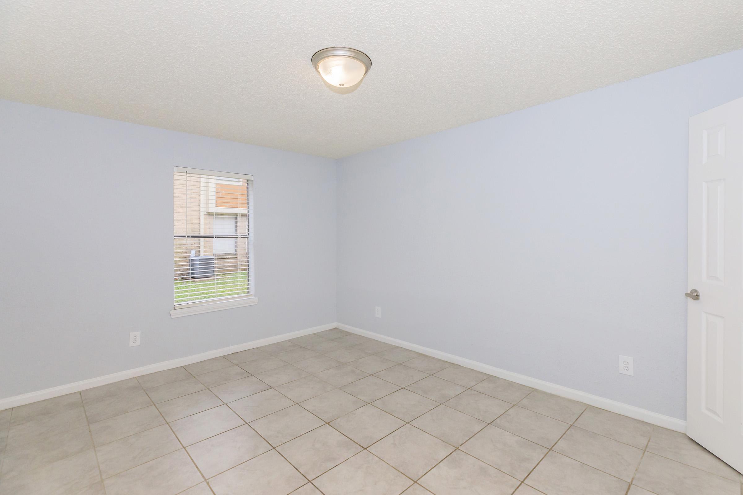 A vacant room with light blue walls and a ceiling light fixture. There is a small window with blinds allowing natural light in, and the floor is tiled. The space appears clean and ready for furnishing. A white door is visible on one side.