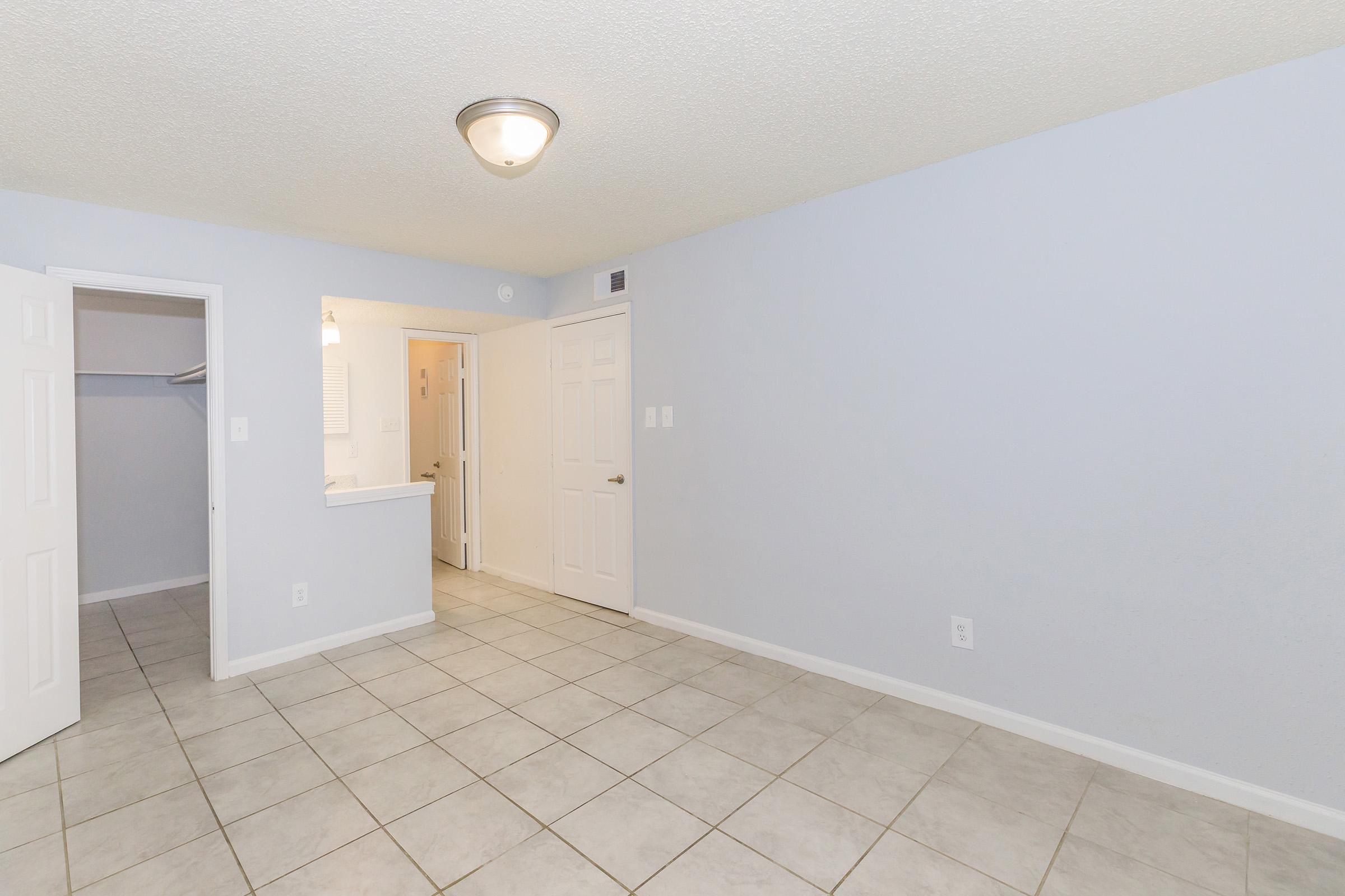 A spacious, empty room with light blue walls and a tiled floor. A ceiling light fixture is visible. The image shows an open doorway leading to another area, possibly a kitchen or bathroom, with a closet space on the left. No furniture or decorations are present, emphasizing the room's openness.
