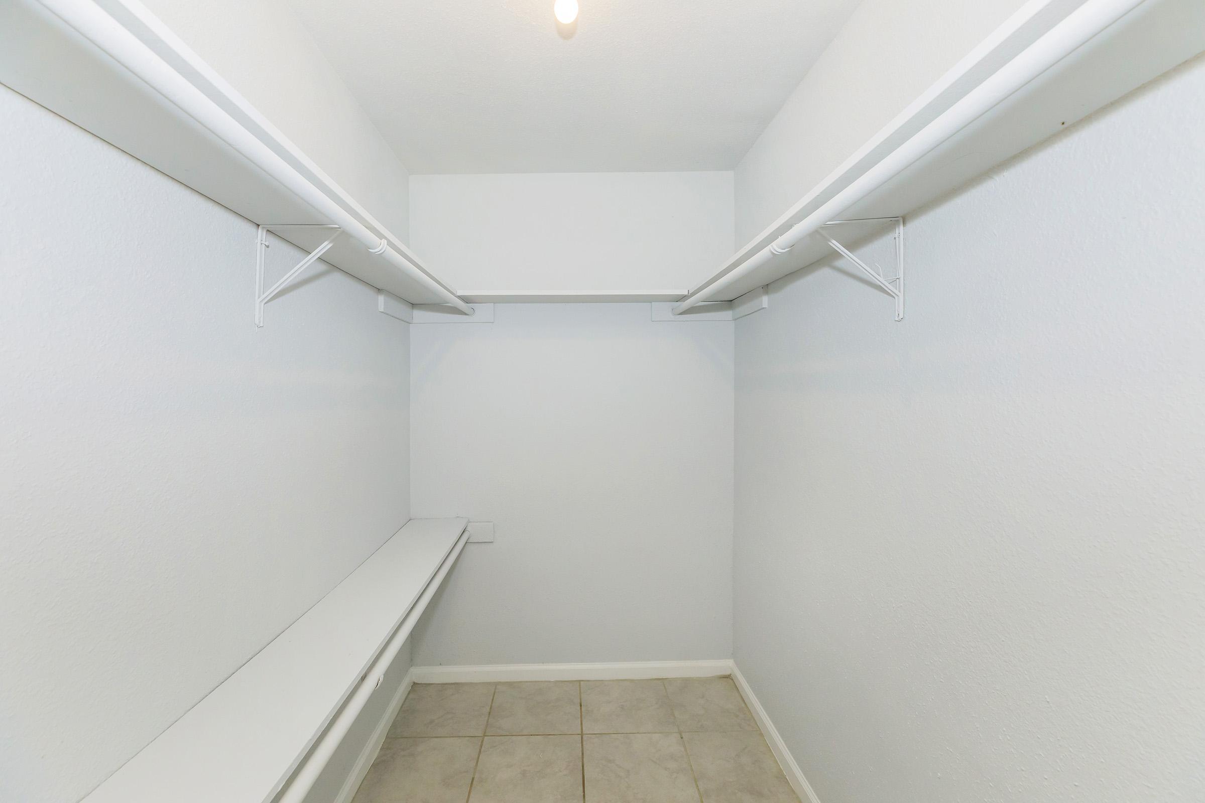 Empty closet interior with white walls and a tile floor. It features two wall-mounted shelves and a long countertop on one side, illuminated by a ceiling light. The space is clean and minimalistic, suitable for storage.