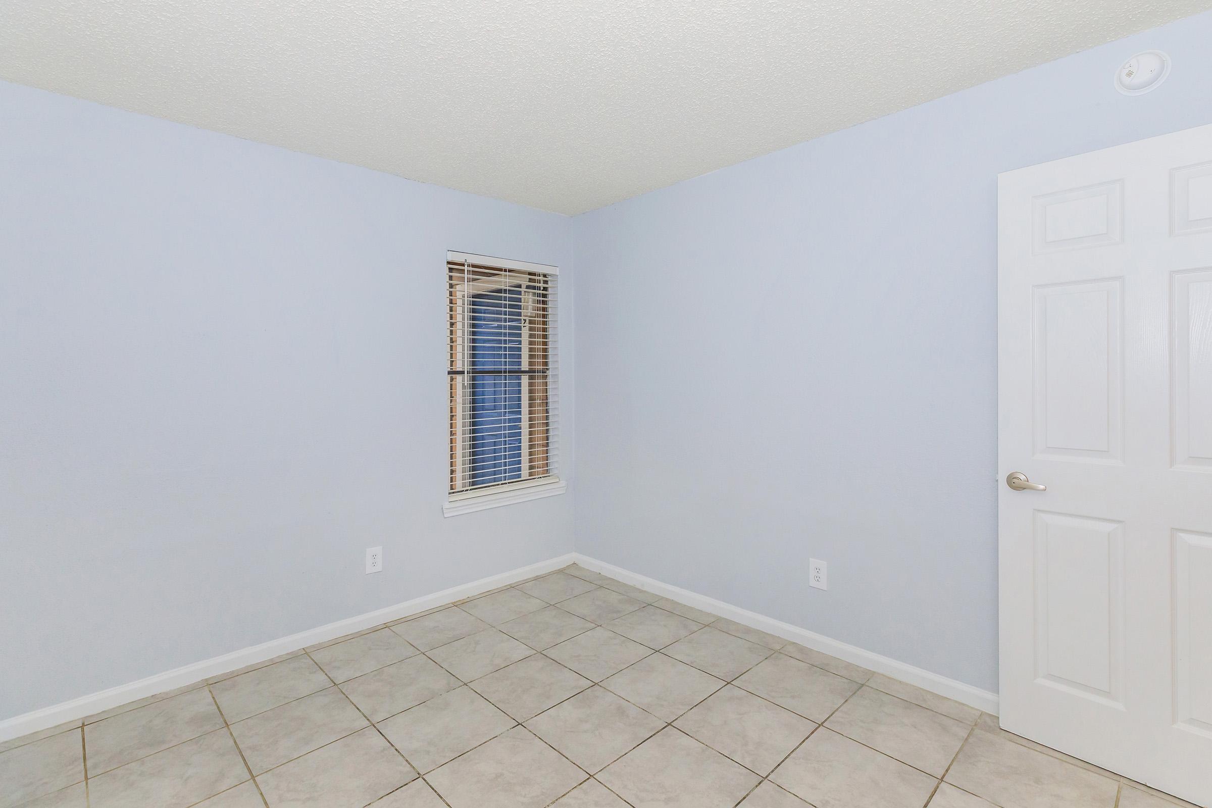A light blue room with a ceiling that has a textured finish. The walls are painted light blue, and there is a small window with white blinds letting in natural light. The flooring is tiled in a neutral color, and a white door is visible on the right side of the image. The room appears empty and unfurnished.