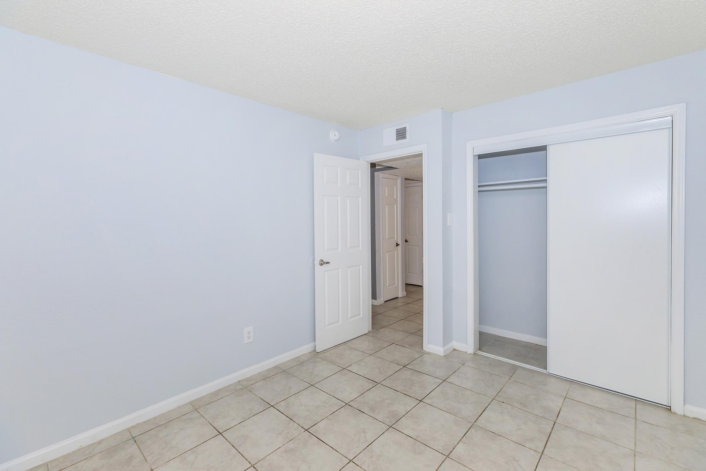 A light blue-painted room featuring tile flooring, a closet with sliding doors, and two white doors leading to other areas. The space is empty and well-lit, with a clean and minimalist aesthetic.