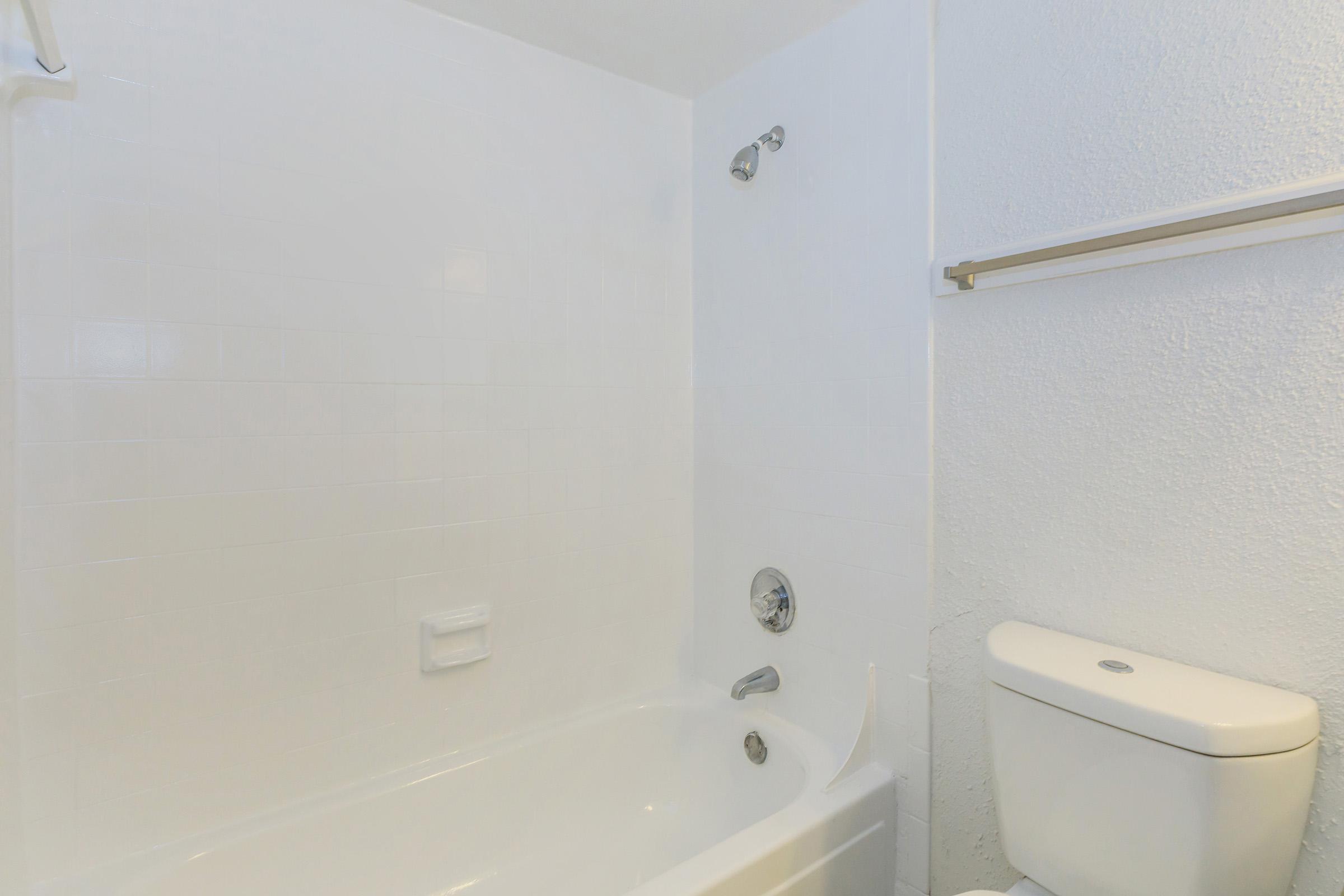 A clean, minimalistic bathroom featuring a bathtub with a showerhead on the wall, a toilet, and white tiled walls. The overall look is bright and modern, with neutral colors creating a simple and uncluttered atmosphere.