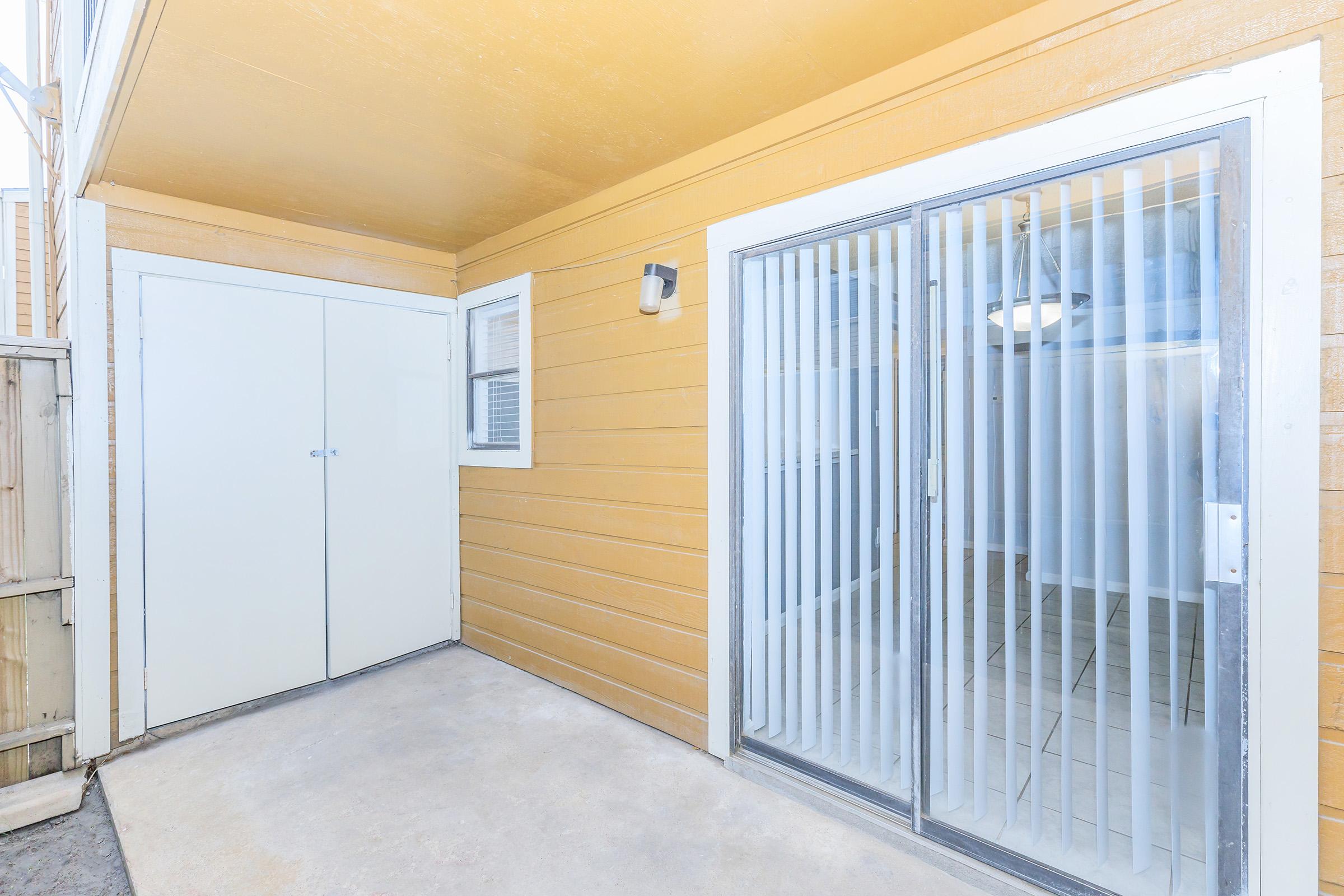 A bright, exterior view of a home featuring a yellow wooden wall, a sliding glass door with vertical blinds, and a small storage area with dual doors. The space is clean and well-lit, showcasing a concrete floor and an inviting entrance.