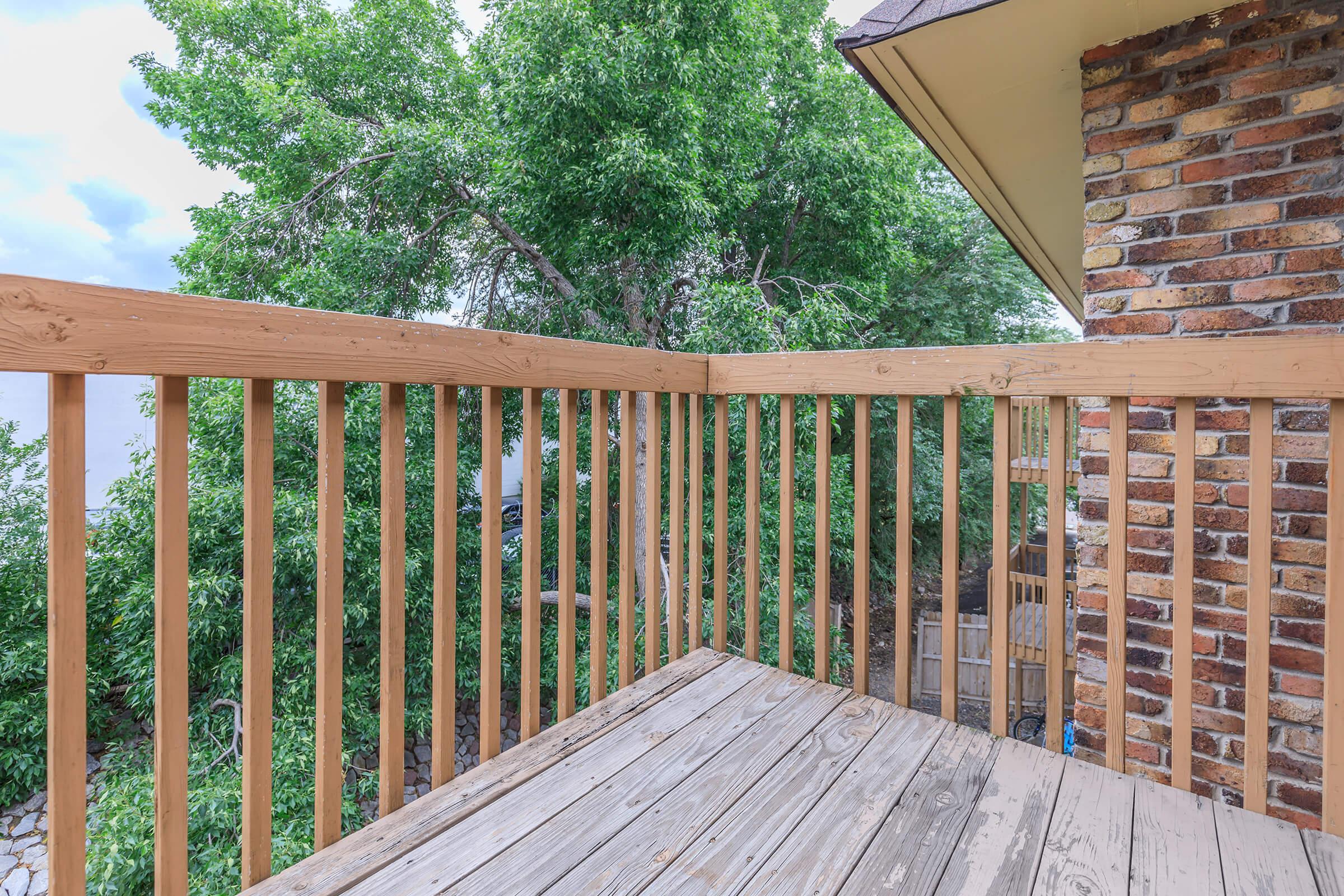 A wooden deck with a railing, surrounded by greenery. The view includes trees and part of a brick building in the background. The scene is bright and natural, suggesting an outdoor space for relaxation.