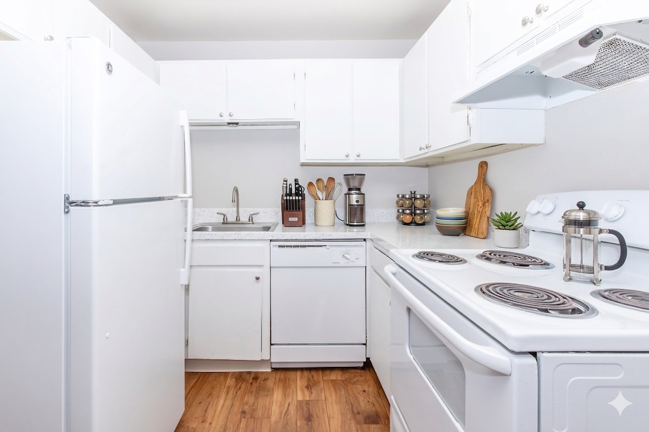 A modern kitchen featuring white cabinets and appliances, including a refrigerator, dishwasher, and stove with an oven. The countertop is equipped with a sink, cutting board, and kitchen utensils. A coffee maker and a small plant are visible, along with wooden accents and warm wood flooring.