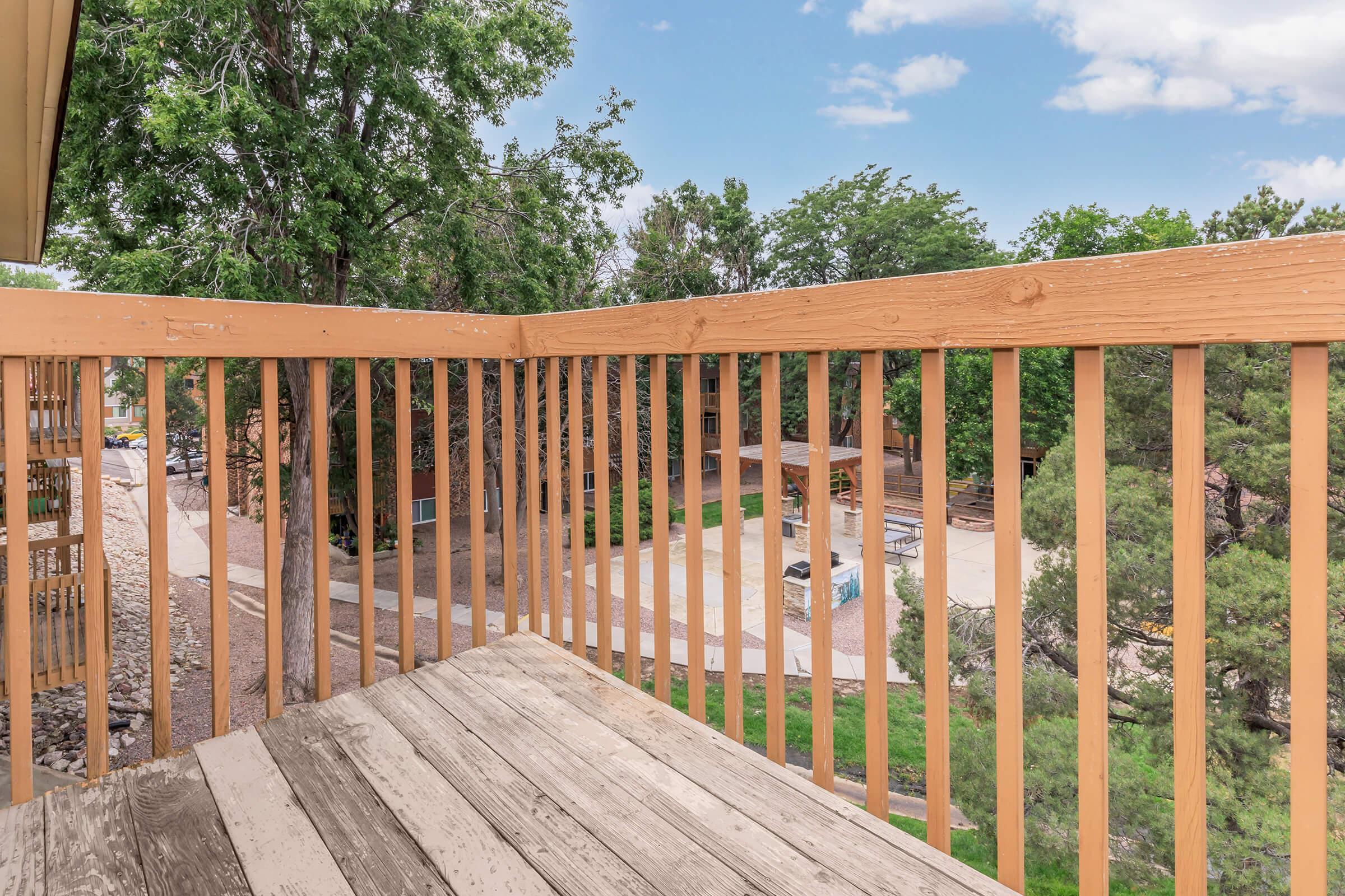 View from a wooden balcony railing overlooking a green landscape with trees. The ground below features a swimming pool and patio area, hinting at residential buildings in the distance. The sky is partly cloudy, creating a relaxed atmosphere.