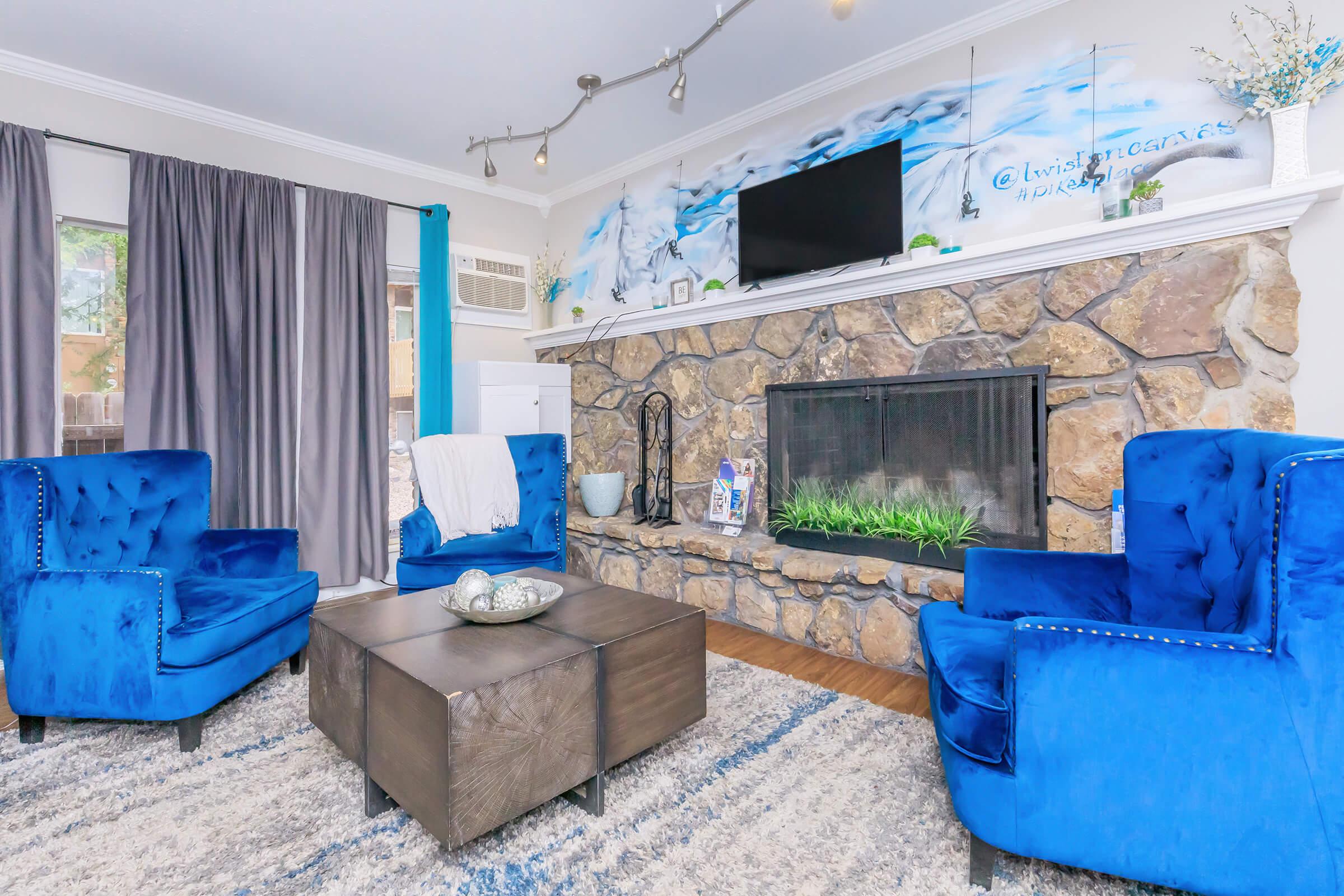 A cozy living room featuring two bright blue armchairs and a dark wooden coffee table. The stone fireplace is adorned with decorative elements, and there's a flat-screen TV mounted above. Natural light filters through the curtains, enhancing the inviting atmosphere.