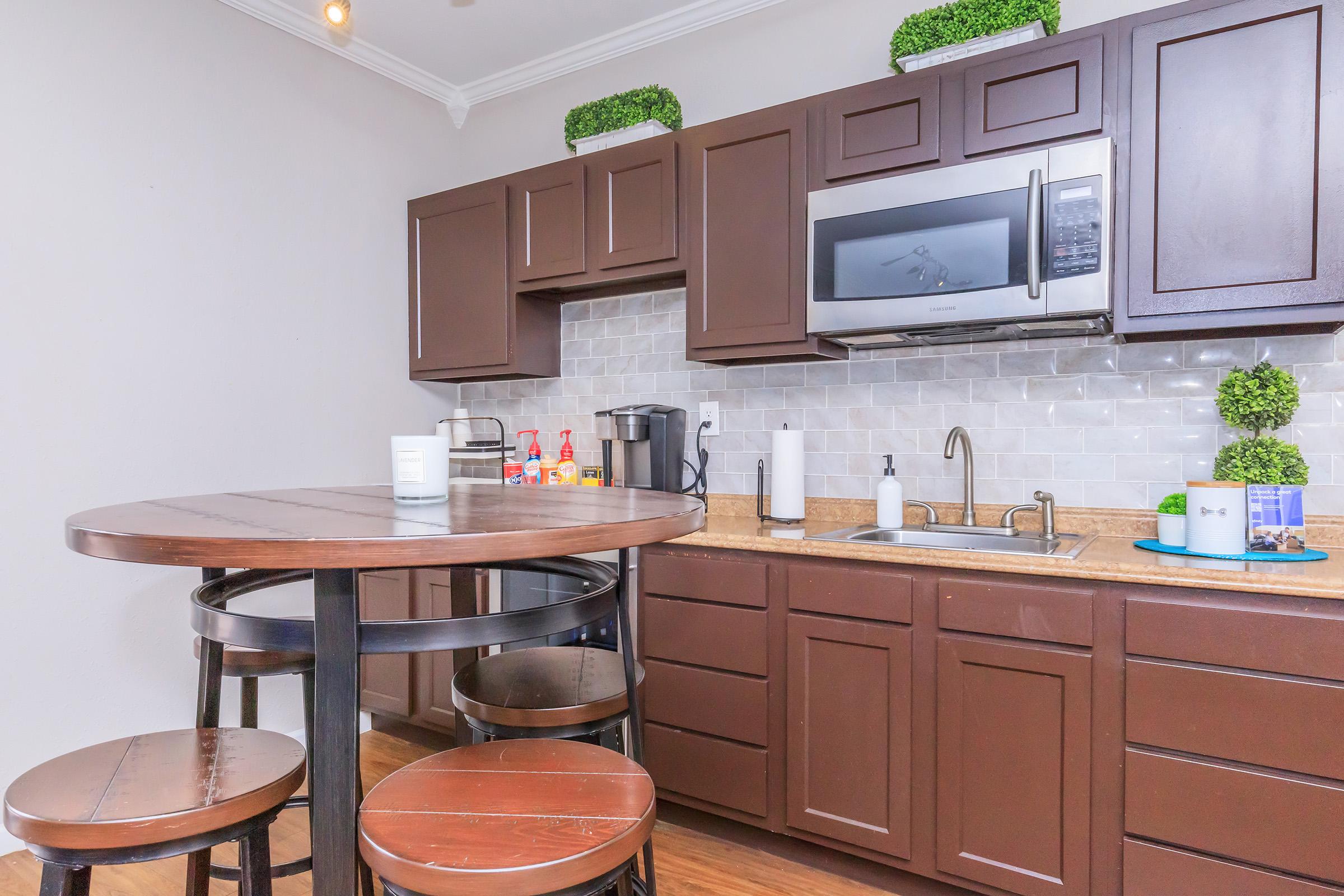 Modern kitchen featuring dark wooden cabinets, a stainless steel microwave, and a countertop with a sink. A round wooden table with three black stools is in the foreground, and small green potted plants are placed on the cabinets above the backsplash of light-colored tiles.