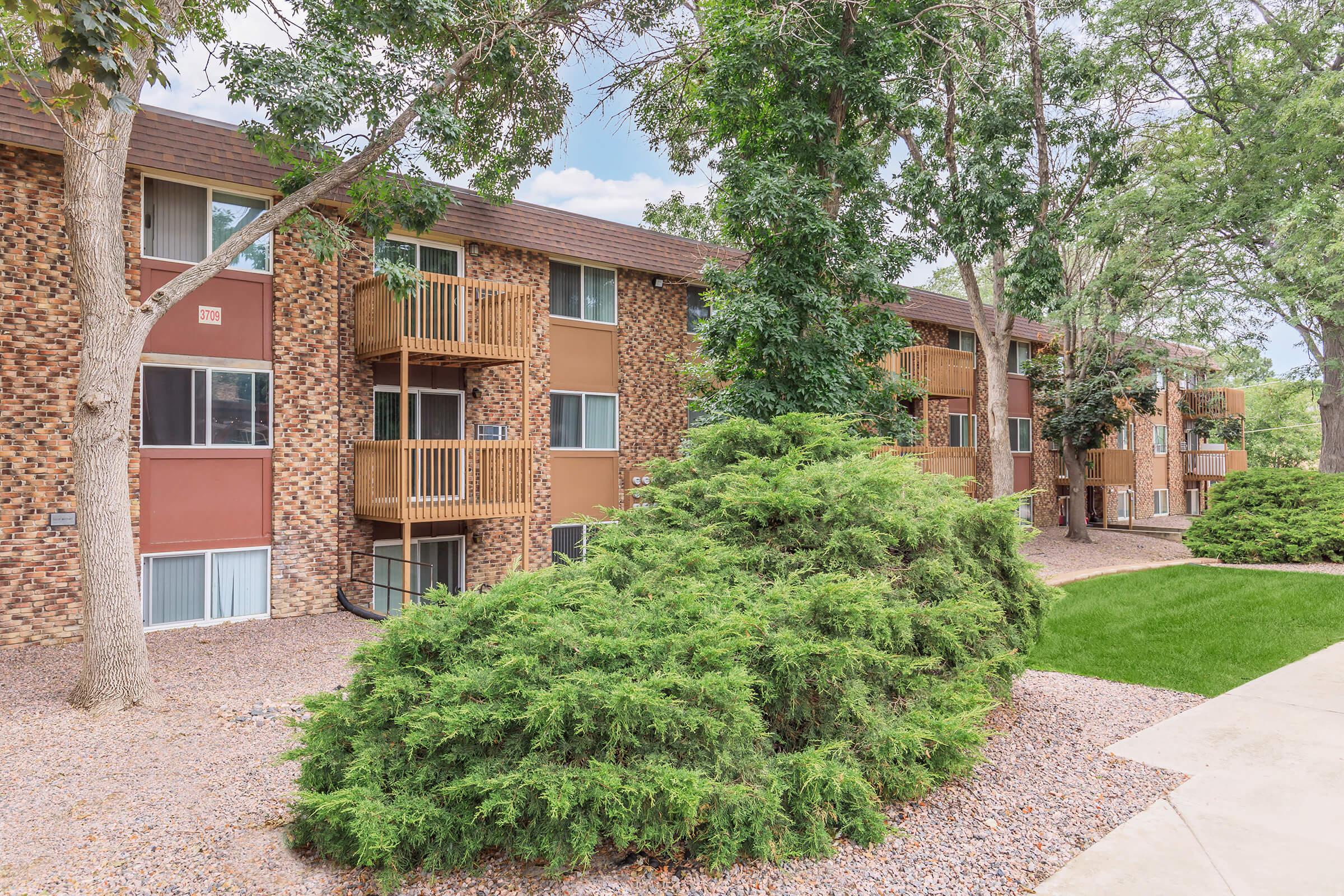 A view of a modern apartment building surrounded by green trees and shrubs. The building features a mix of brick and tan siding, with several balconies visible. The landscaping includes well-maintained grass and a pathway leading up to the entrance.
