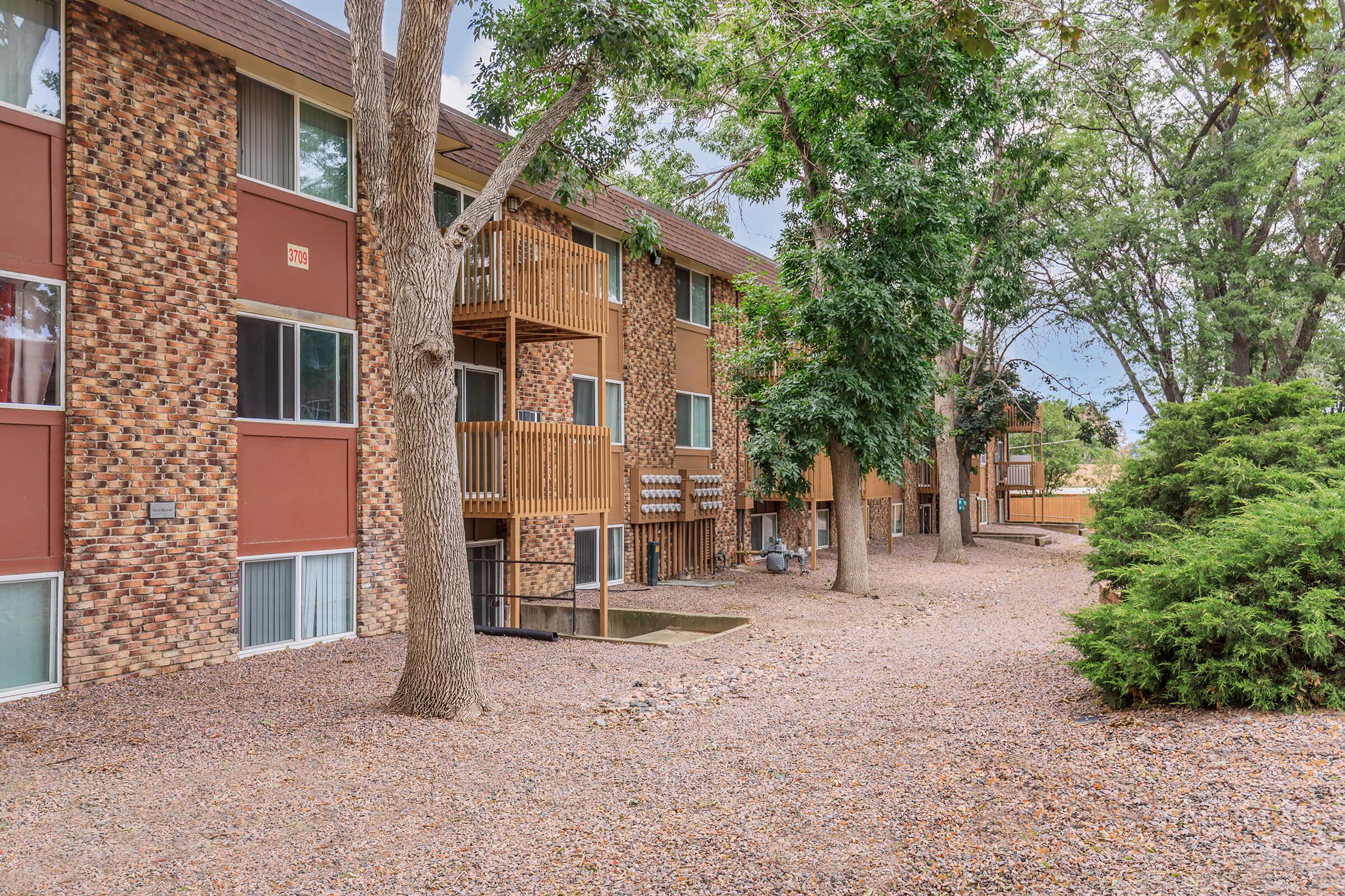 A view of an apartment complex surrounded by trees, with a gravel pathway leading to the building. The structure features brick and beige siding, with balconies visible on the upper floors. Lush greenery and a well-maintained landscape complement the setting, suggesting a tranquil residential environment.