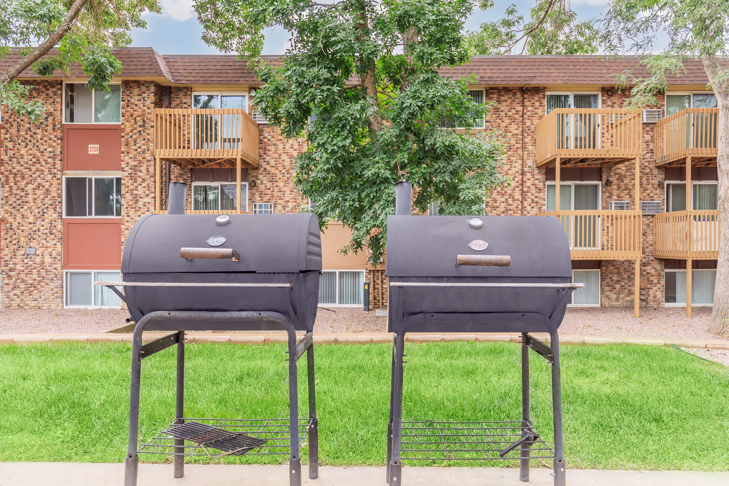 Two black charcoal grills stand on metal stands in a grassy area, with a brick apartment building in the background. The building features wooden balconies and is surrounded by trees.