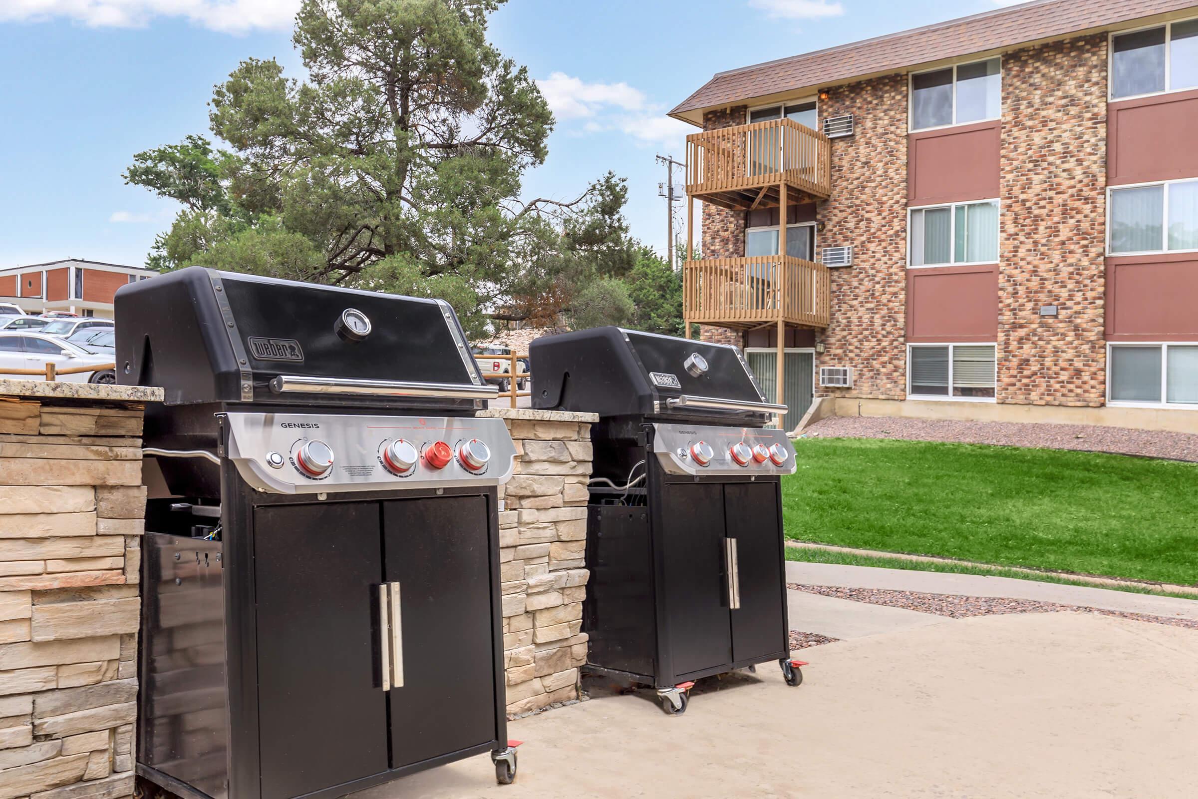 Two black gas grills with stainless steel accents are positioned on a stone patio. In the background, there is a multi-story apartment building with brick and siding exteriors, surrounded by green grass and trees, indicating a residential outdoor area.