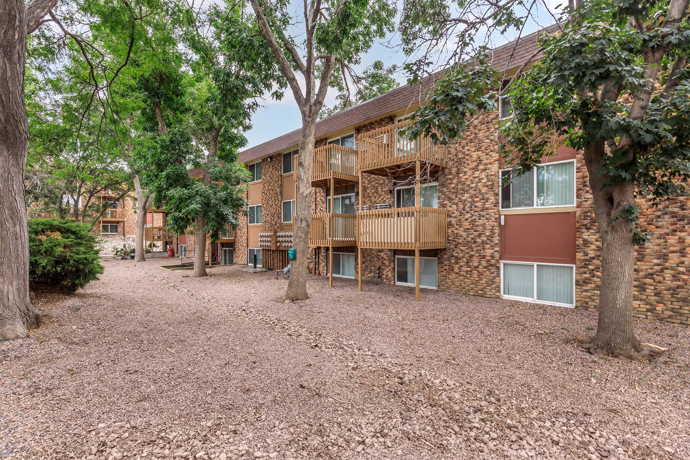 A view of a multi-unit residential building featuring multiple balconies, surrounded by trees and gravel landscaping. The building has a mix of brick and stucco exteriors, with several windows and a serene outdoor area.