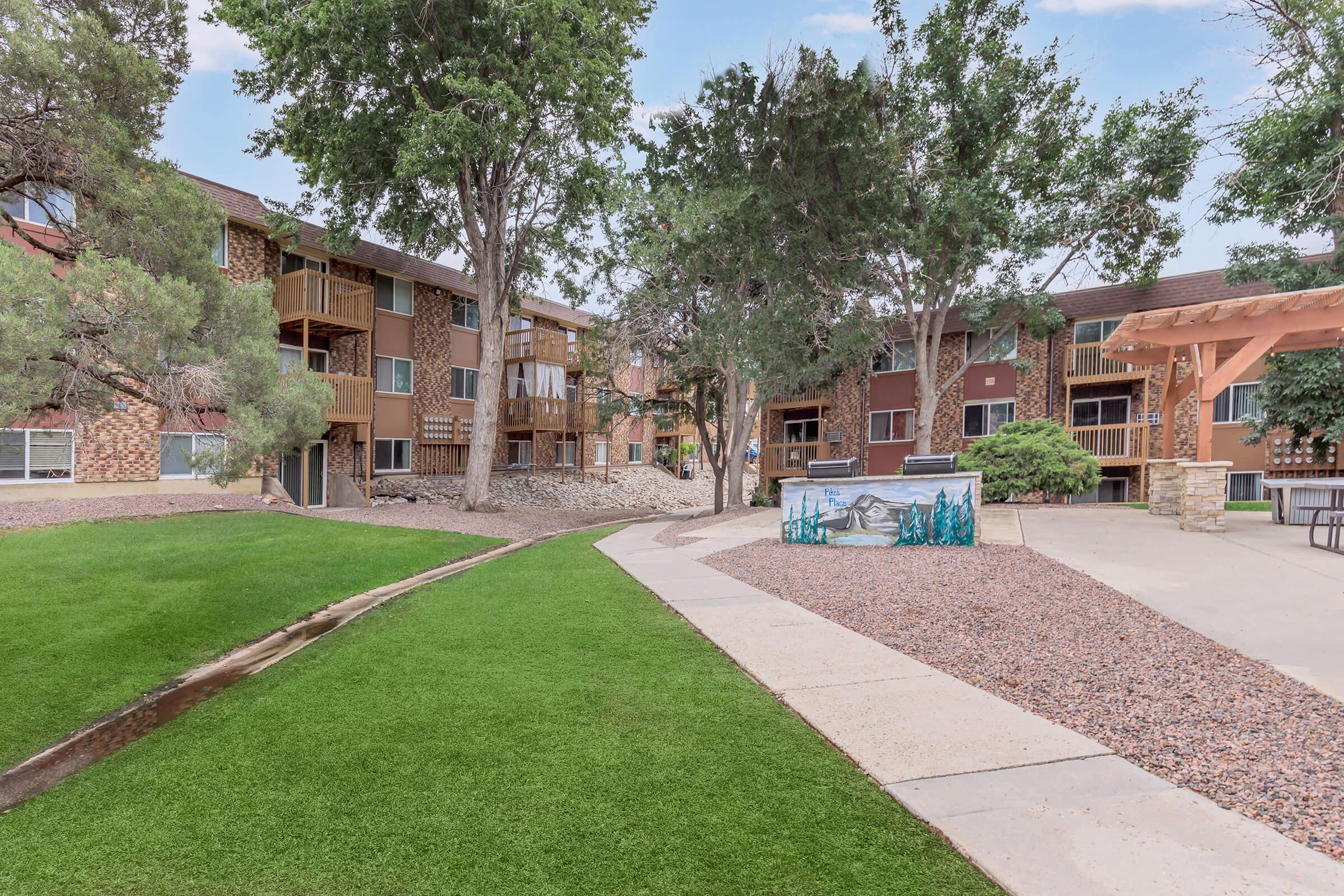 A landscaped courtyard featuring manicured green lawn, surrounded by multi-story apartment buildings. The area includes trees, gravel paths, and a decorative mural. The setting suggests a serene residential community with outdoor space for relaxation and social activities.