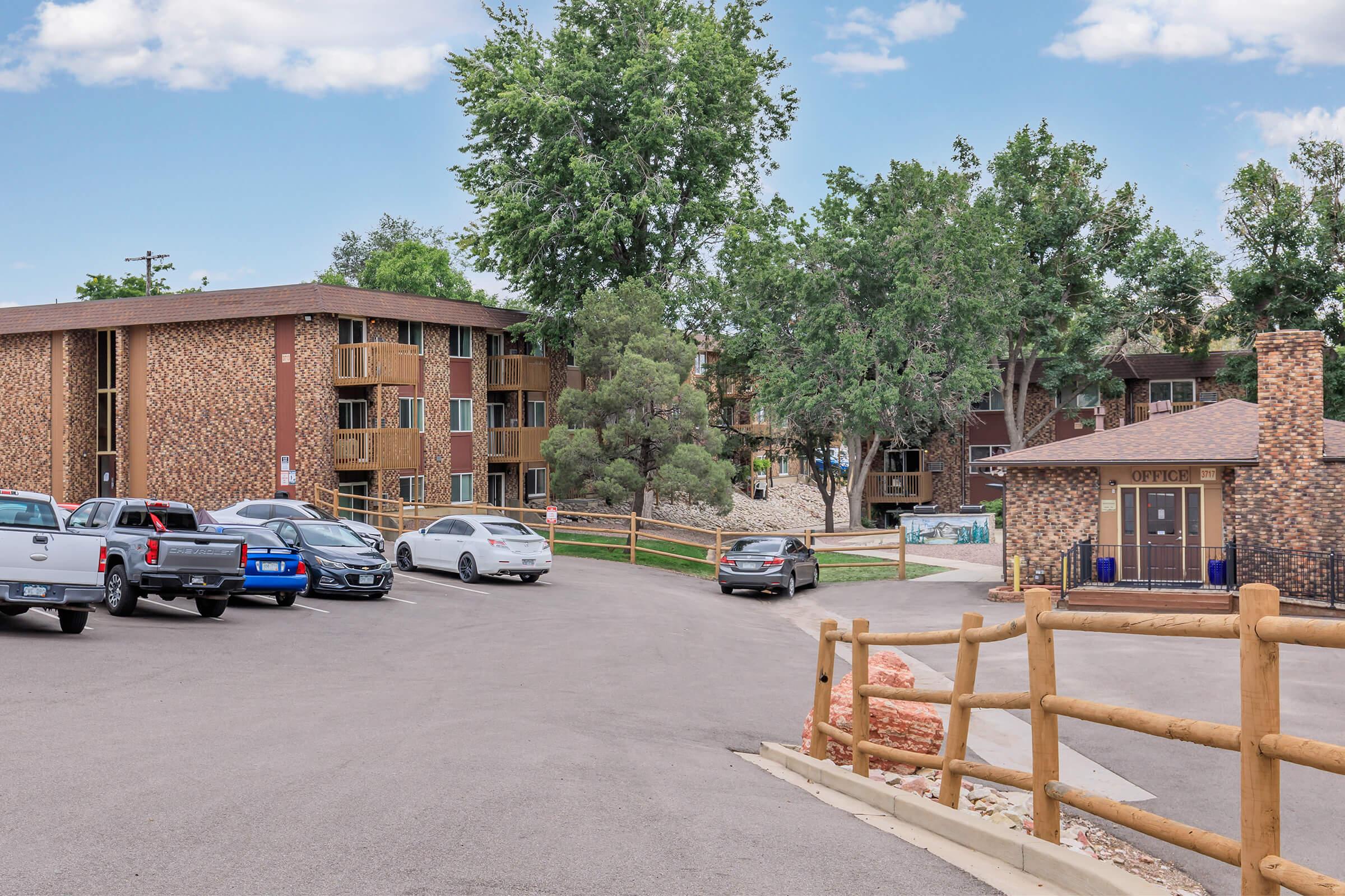View of an apartment complex with two multi-story buildings featuring brick-like facades, surrounded by trees and landscaping. A parking lot is visible with several cars parked, and a small office building marked "OFFICE" is situated in the foreground, along with a wooden fence lining the road.