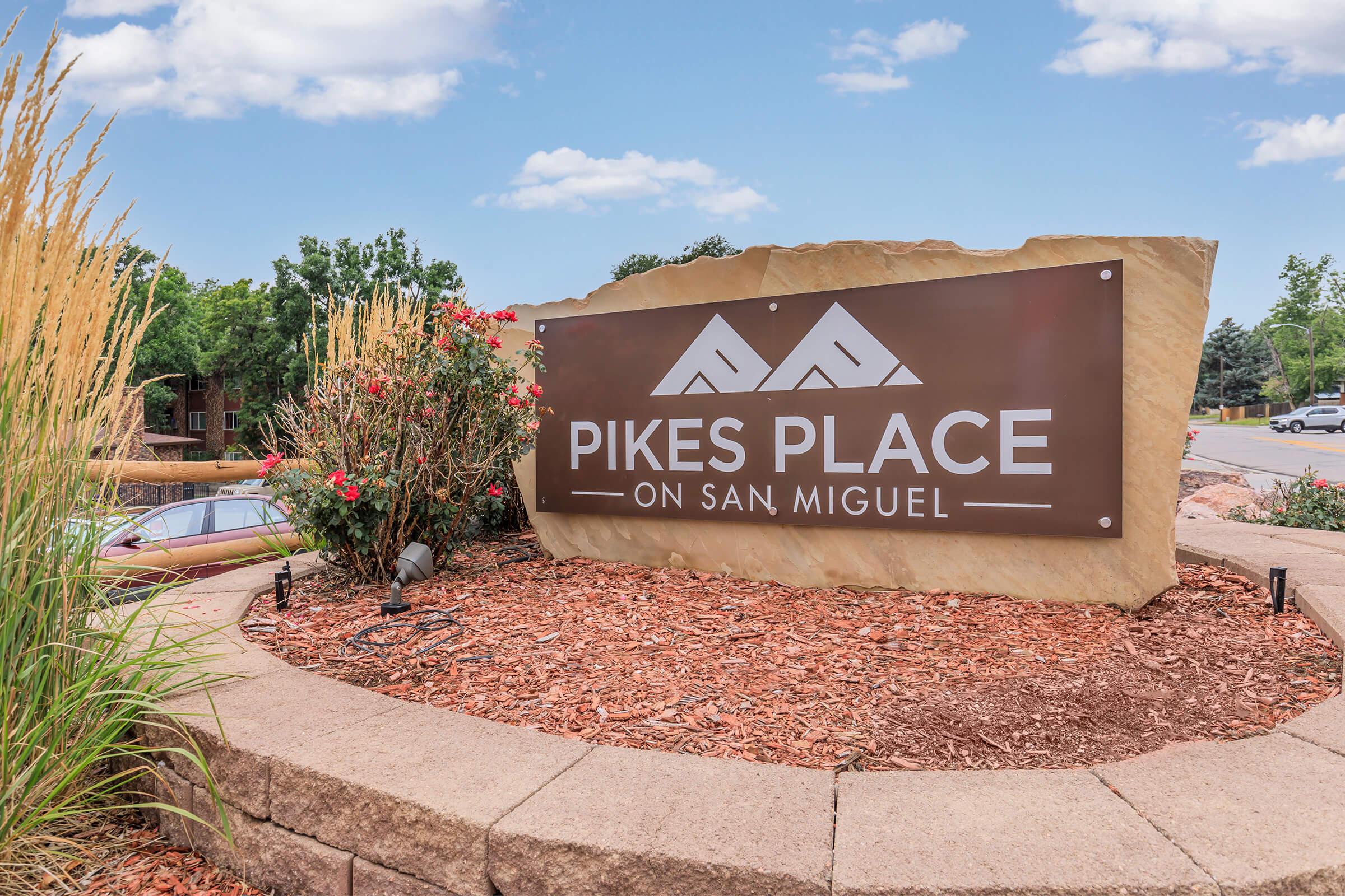 A stone sign displaying "Pikes Place on San Miguel," surrounded by colorful flowers and grasses, set against a blue sky with wispy clouds. The sign features a mountain design and is positioned in a landscaped area.
