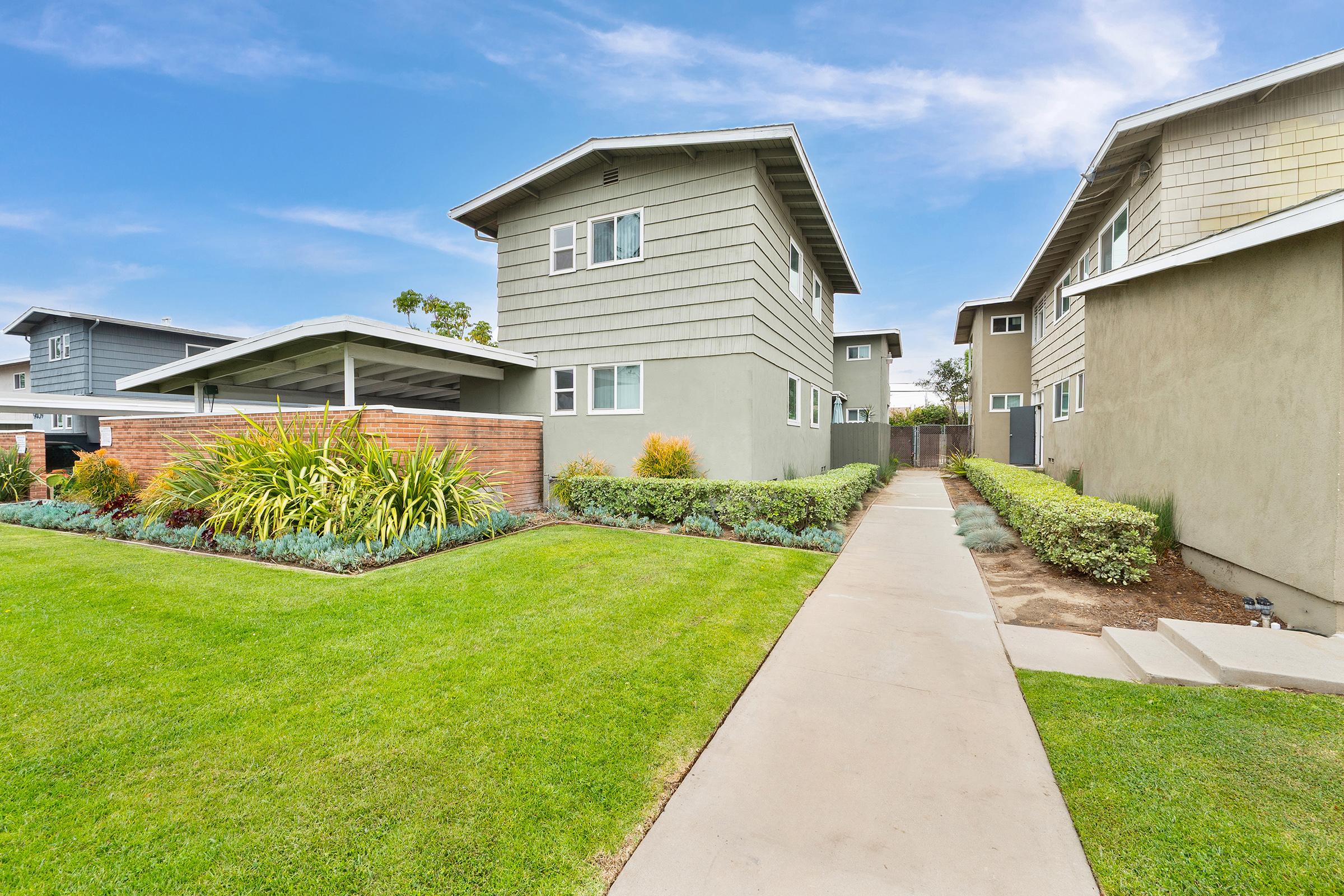 View of a modern residential complex featuring two-story buildings with a mix of gray and green siding. A well-maintained pathway leads to the entrance, bordered by lush greenery and landscaped gardens under a clear blue sky. The neatly trimmed lawn adds to the inviting appearance of the property.