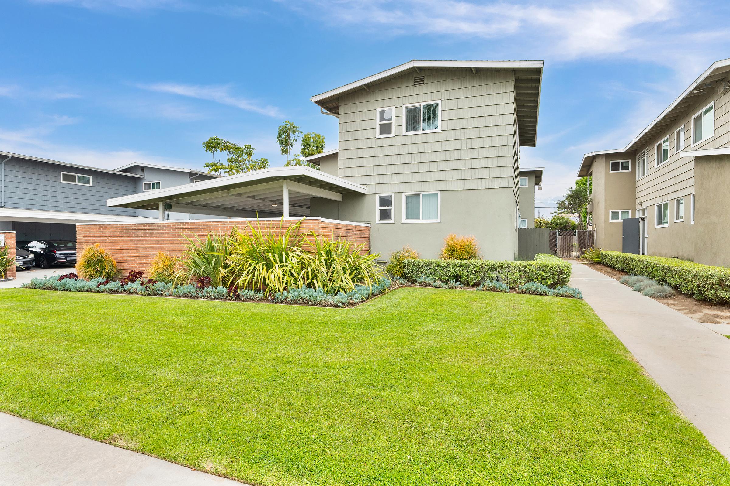 A modern two-story house with a gray exterior, surrounded by well-maintained green lawns and landscaping. In the foreground, a paved walkway leads to the entrance, and a carport is visible. The background features additional residential buildings, creating a suburban setting.
