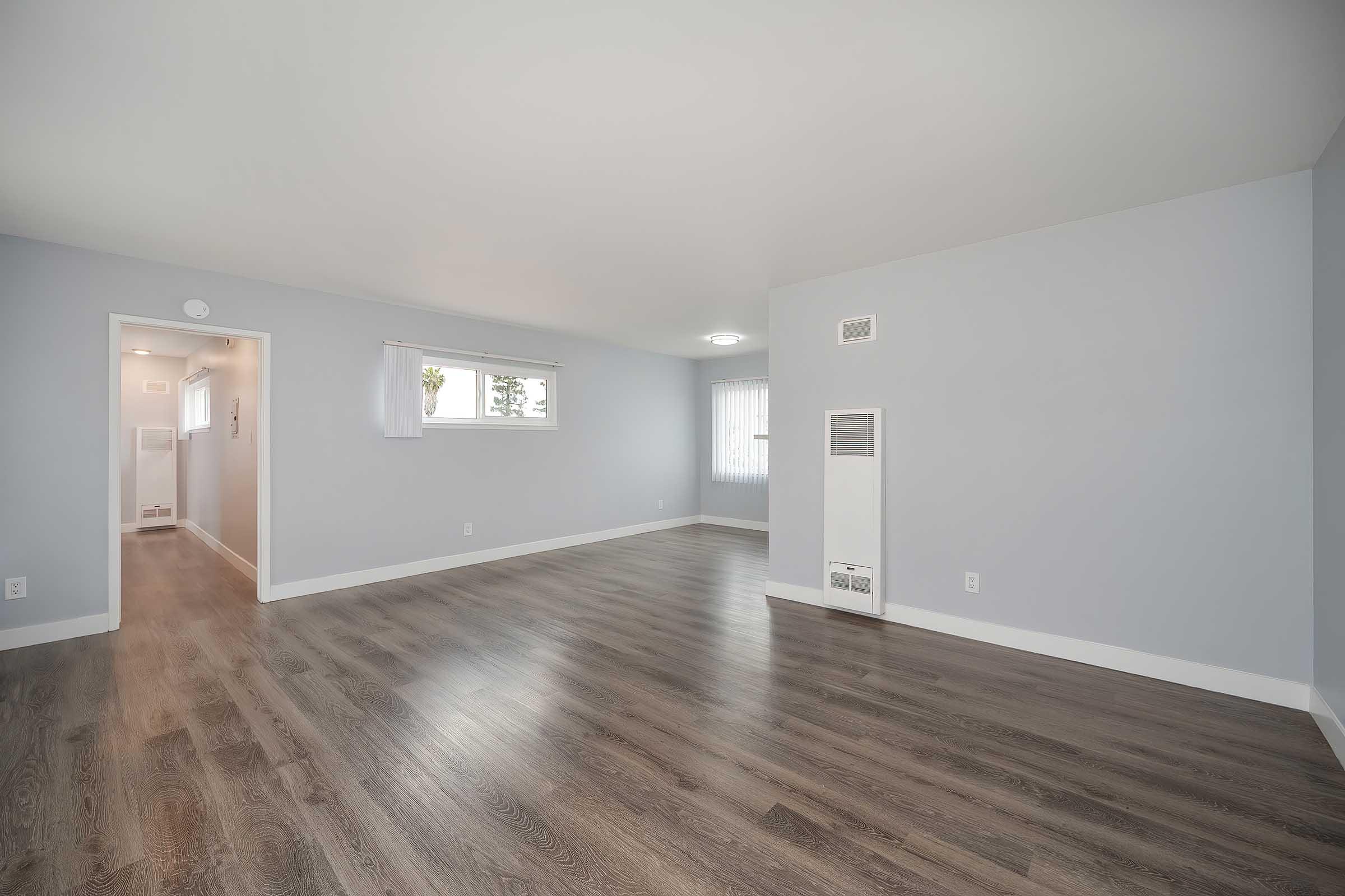 A spacious, empty room with light gray walls and laminate flooring. Natural light filters in through a window, and there is a wall-mounted heater. A doorway leads to another part of the home. The overall atmosphere is bright and inviting, suitable for various furnishings and decor.