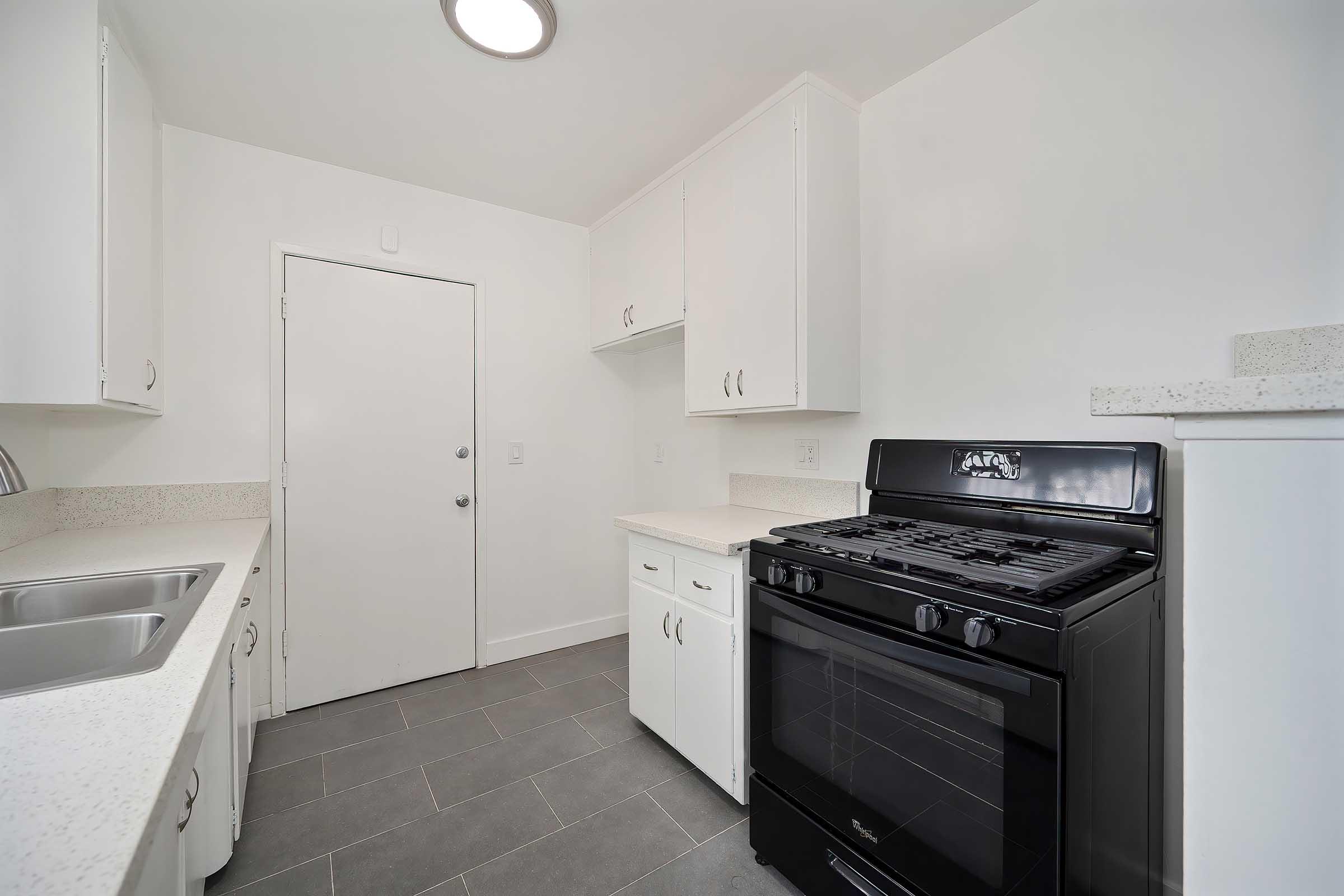A modern kitchen featuring white cabinetry, a black gas stove, and countertop. There are dual sinks and a light-colored countertop. A door leads to an exterior space, and the flooring is gray tiles, contributing to a clean and contemporary design.