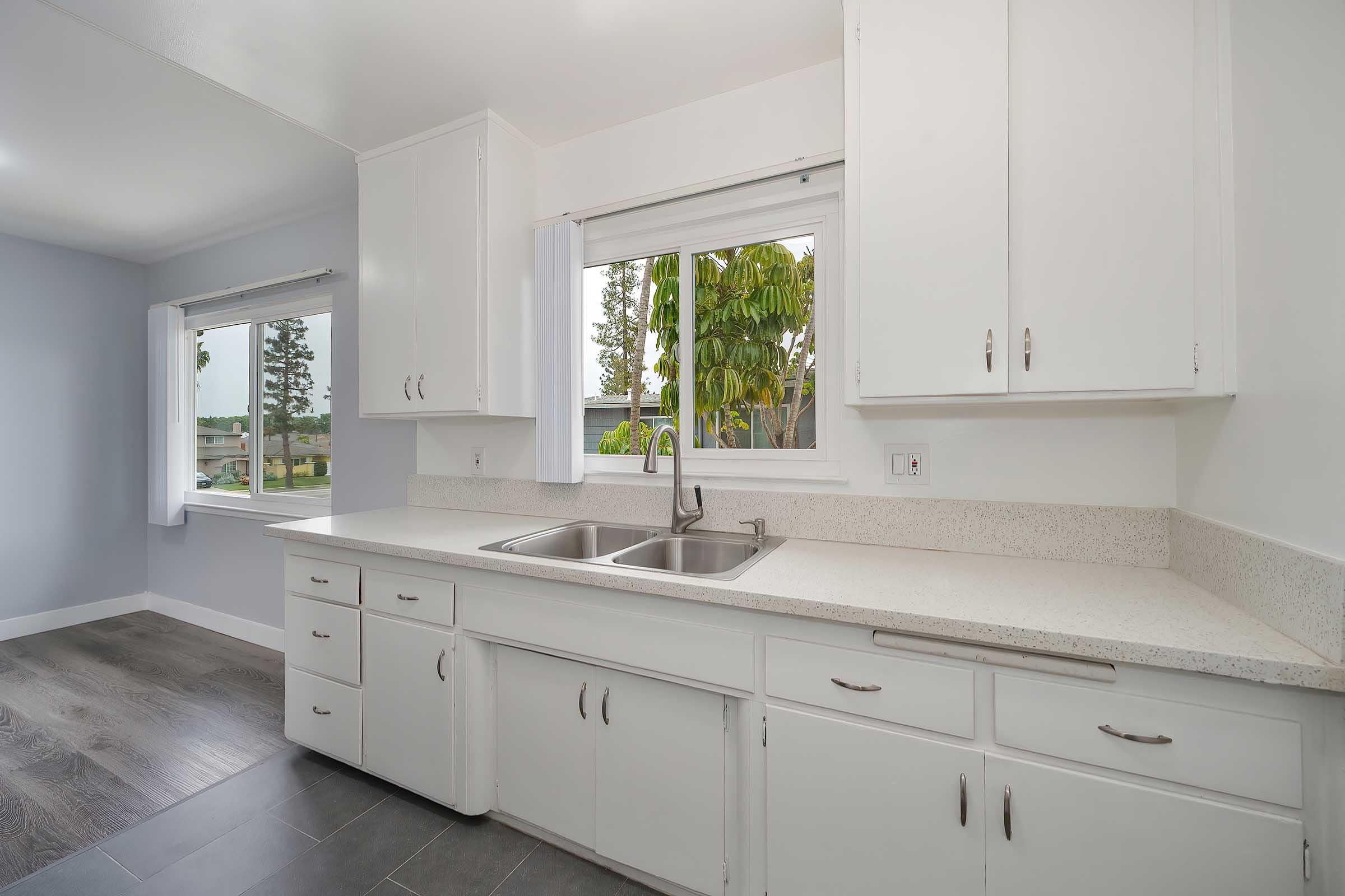 Bright kitchen with white cabinets and a double sink under a window. Light-colored countertops and gray flooring give a modern feel. Natural light streams in from the window, which offers a view of greenery outside. The room has a clean, open layout with plenty of storage space.