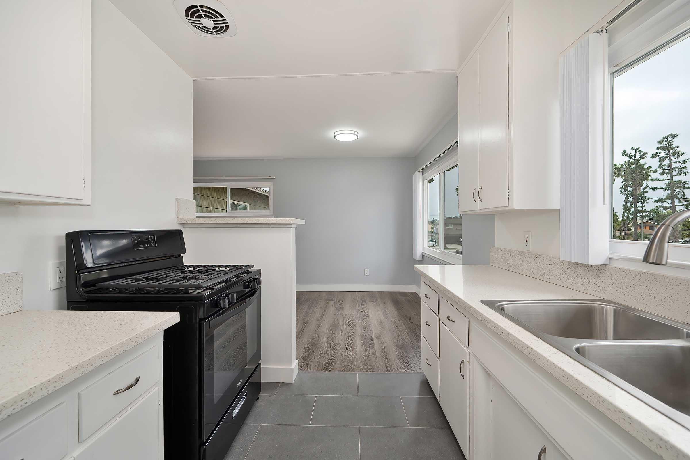 A modern kitchen featuring a black gas stove, white cabinets, and a dual sink. The countertops are light-colored, and there is a large window allowing natural light. Light gray walls and a wood-style floor add a contemporary touch to the space.