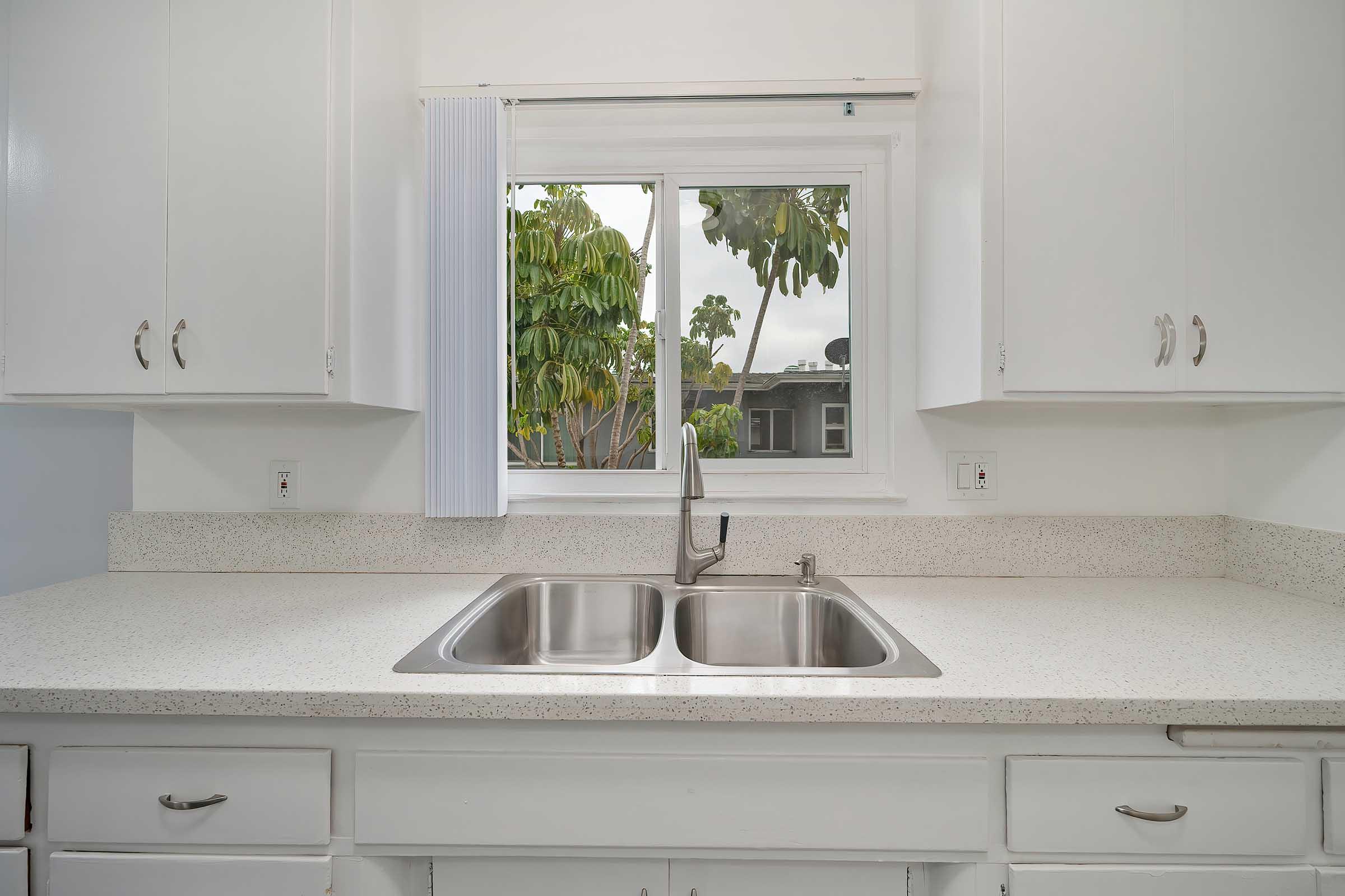 A clean, modern kitchen featuring a double sink with a sleek faucet. The countertop is light-colored and smooth, while white cabinets with silver handles line the space. A window above the sink showcases greenery outside, allowing natural light to brighten the room.
