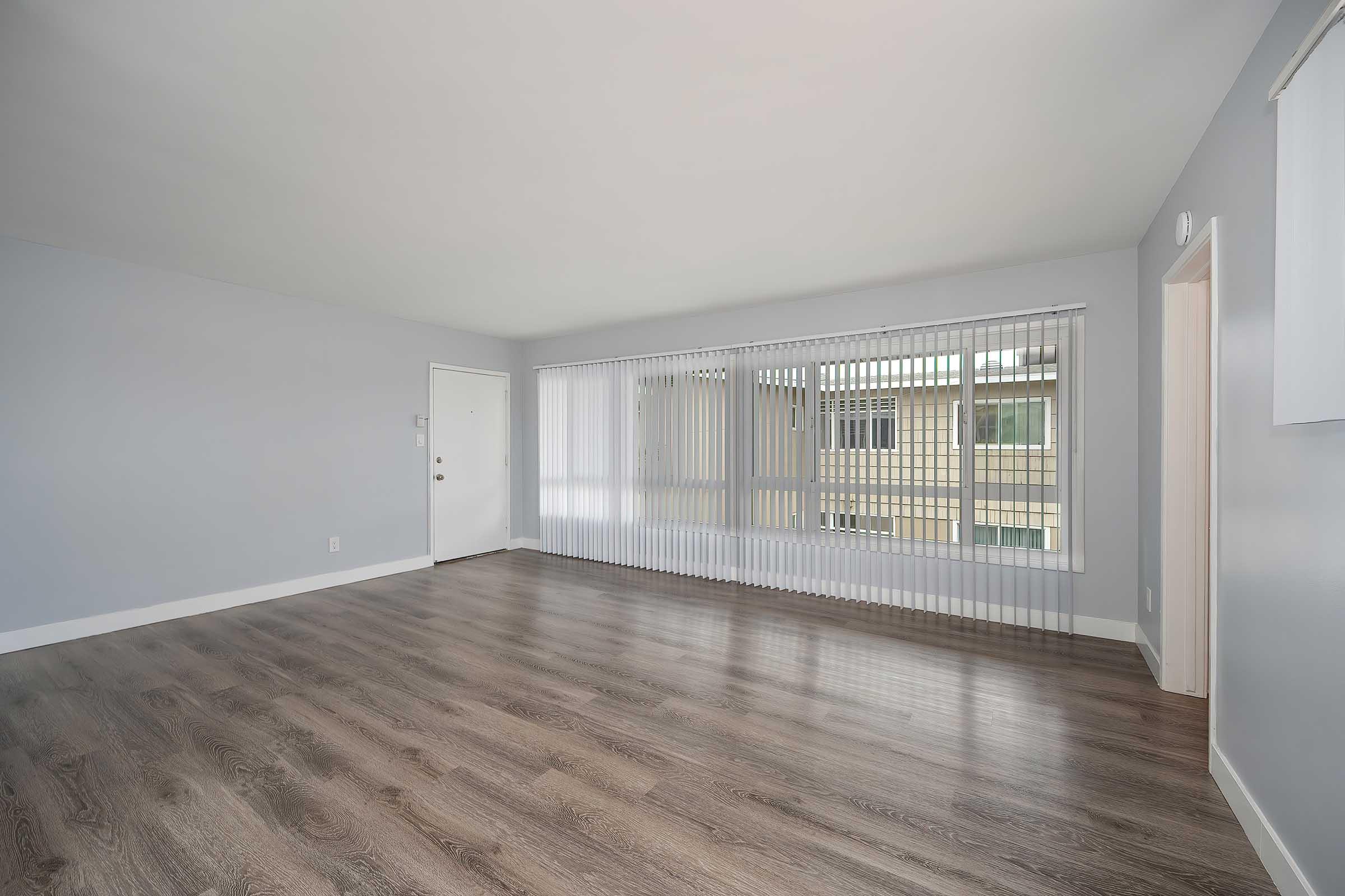 Empty room with light gray walls and a large window covered by vertical blinds. The floor is a medium-tone wood laminate. A door is visible on the left side, providing a sense of natural light and openness. The space is versatile and ready for furniture arrangement.