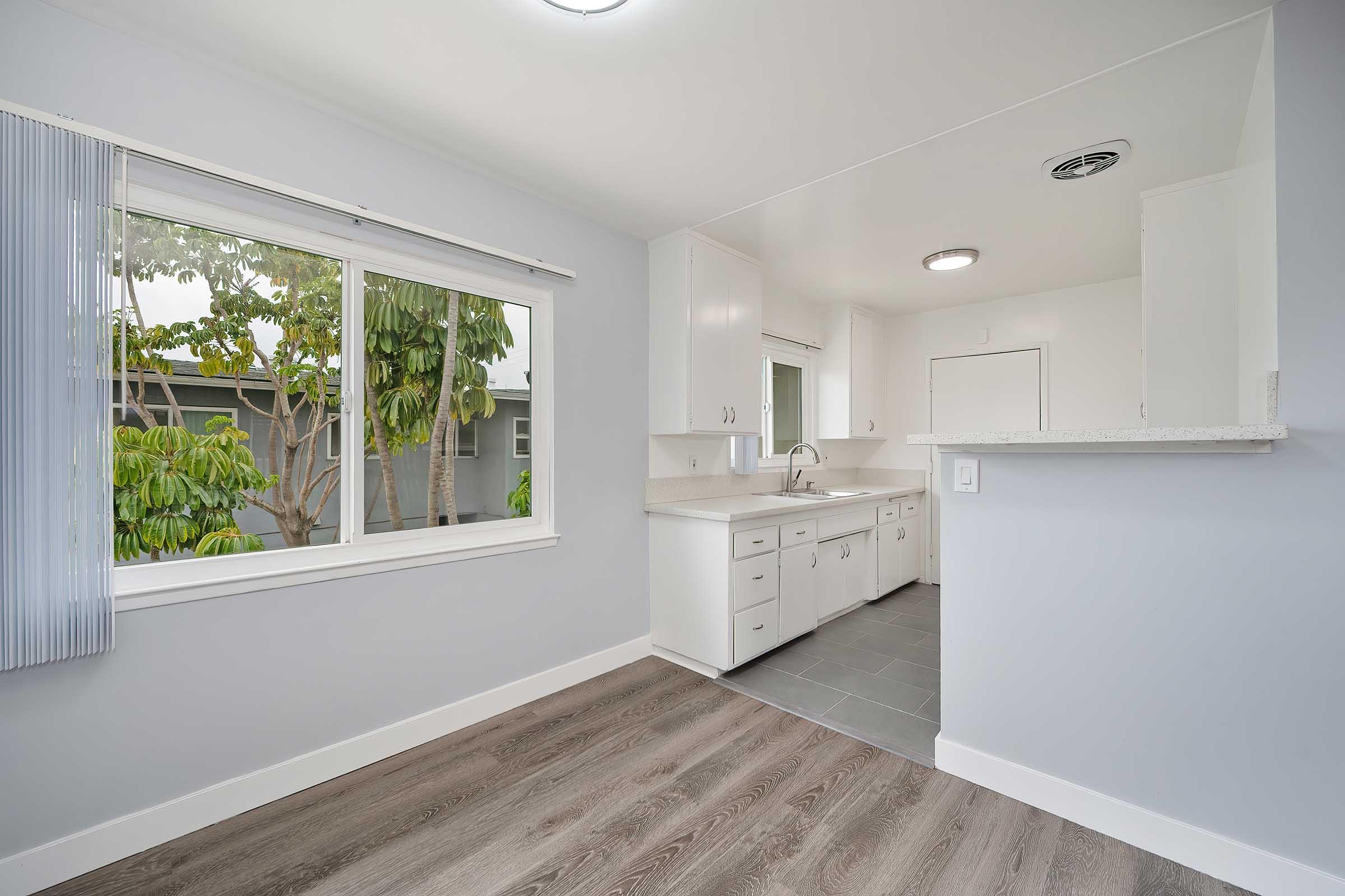 A modern kitchen with white cabinetry and a light gray wall. Natural light streams through a large window, illuminating the space. The flooring is a wood-like laminate, and the kitchen features a sink and countertops. A small entry point leads to another area of the home.