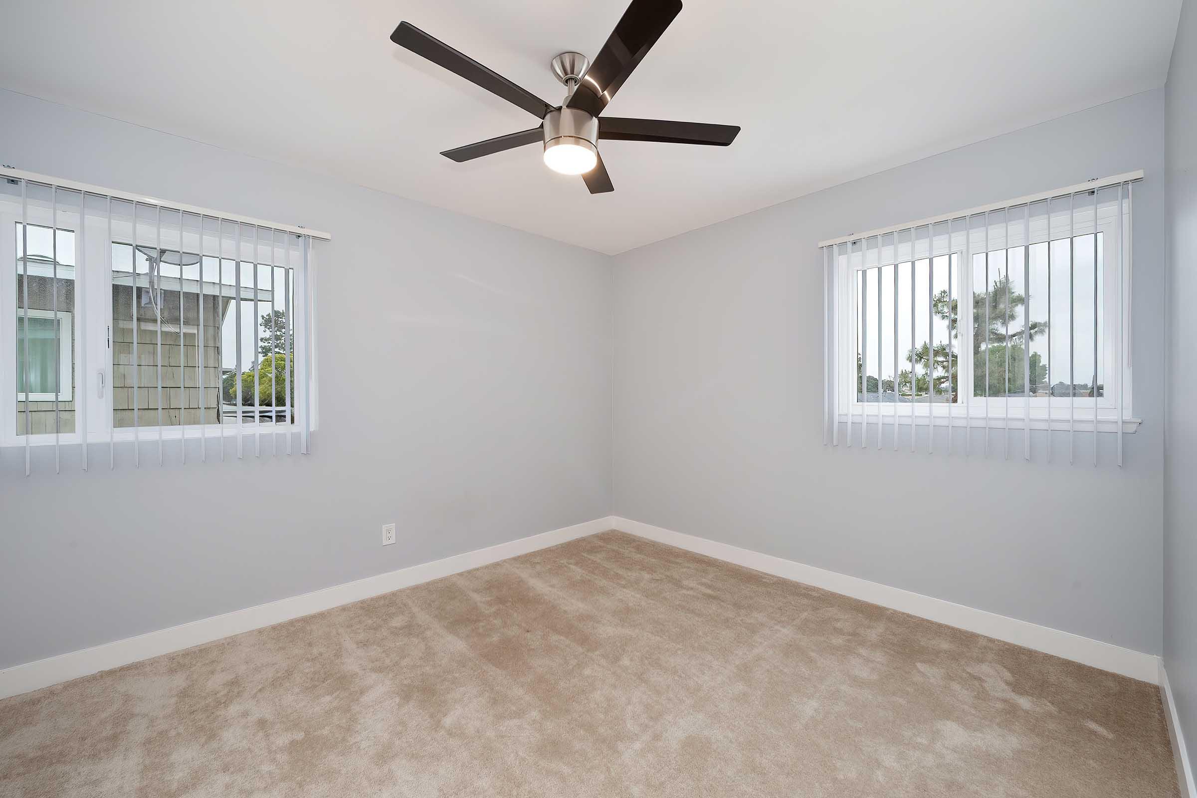 Empty room with light gray walls and a carpeted floor. Two windows with vertical blinds provide natural light. A modern ceiling fan with five blades is mounted in the center, adding a contemporary touch to the space. Ideal for various uses such as a bedroom or office.